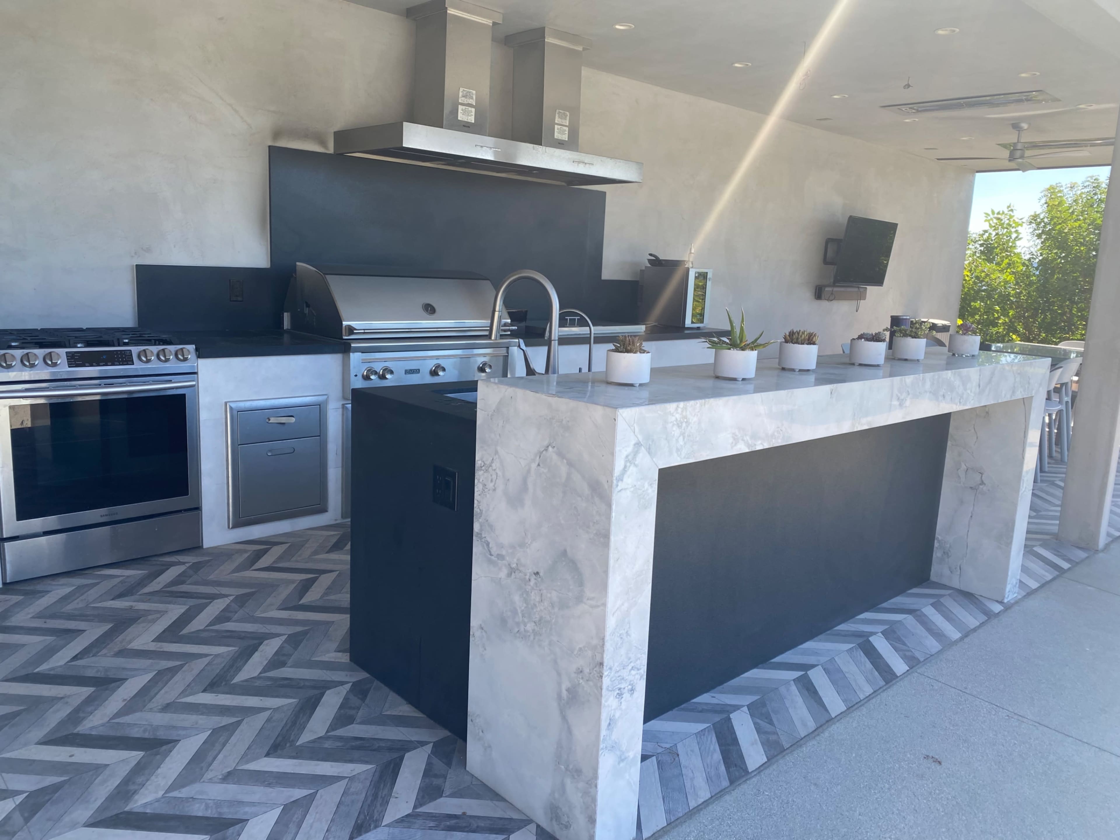 The image shows a modern outdoor kitchen featuring a large stone island, stainless steel appliances, and a chevron-patterned floor.