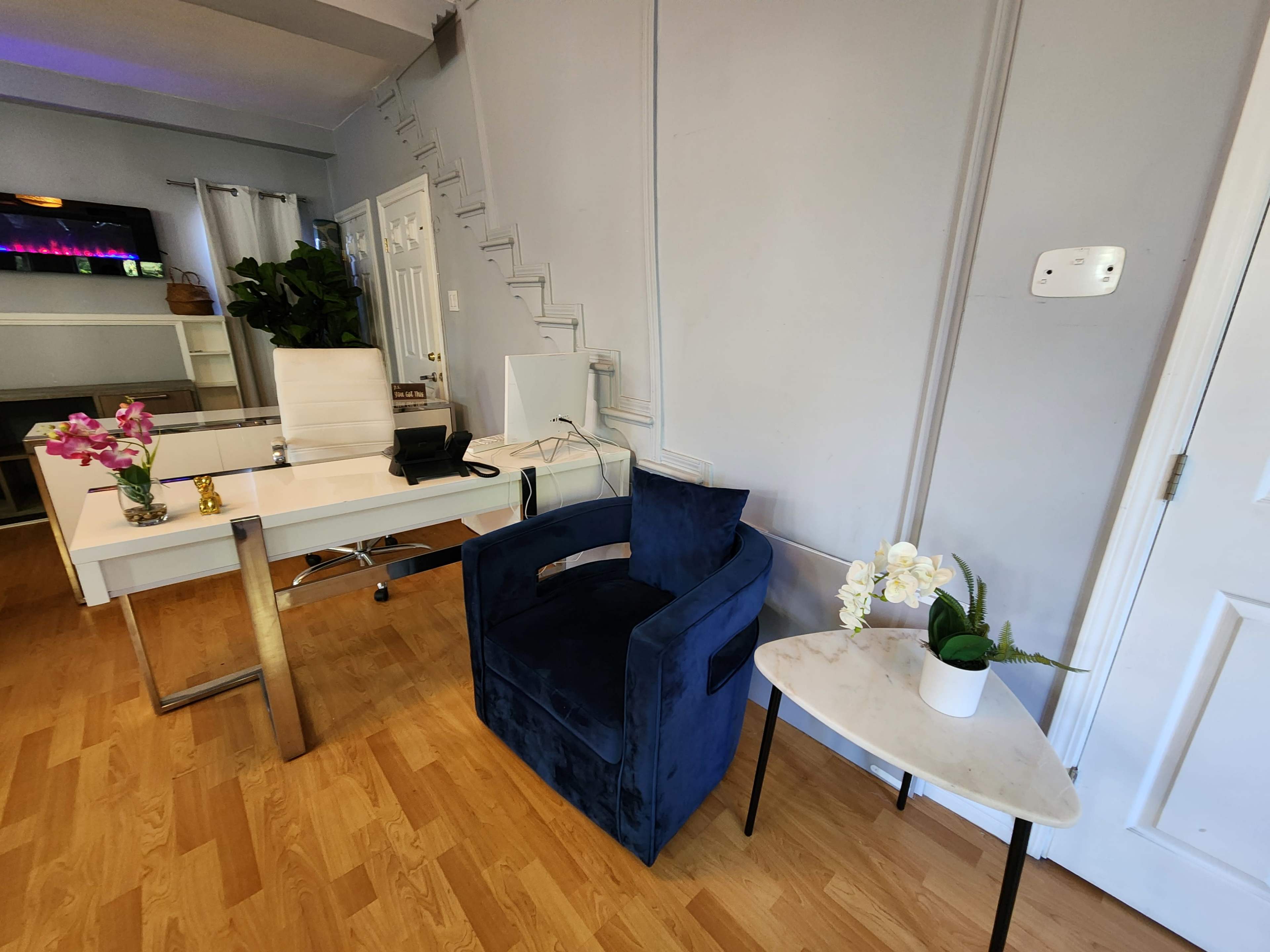 The image shows a modern interior space featuring a blue velvet chair, a small marble side table with a plant, and a white desk with a computer and decorative flowers.