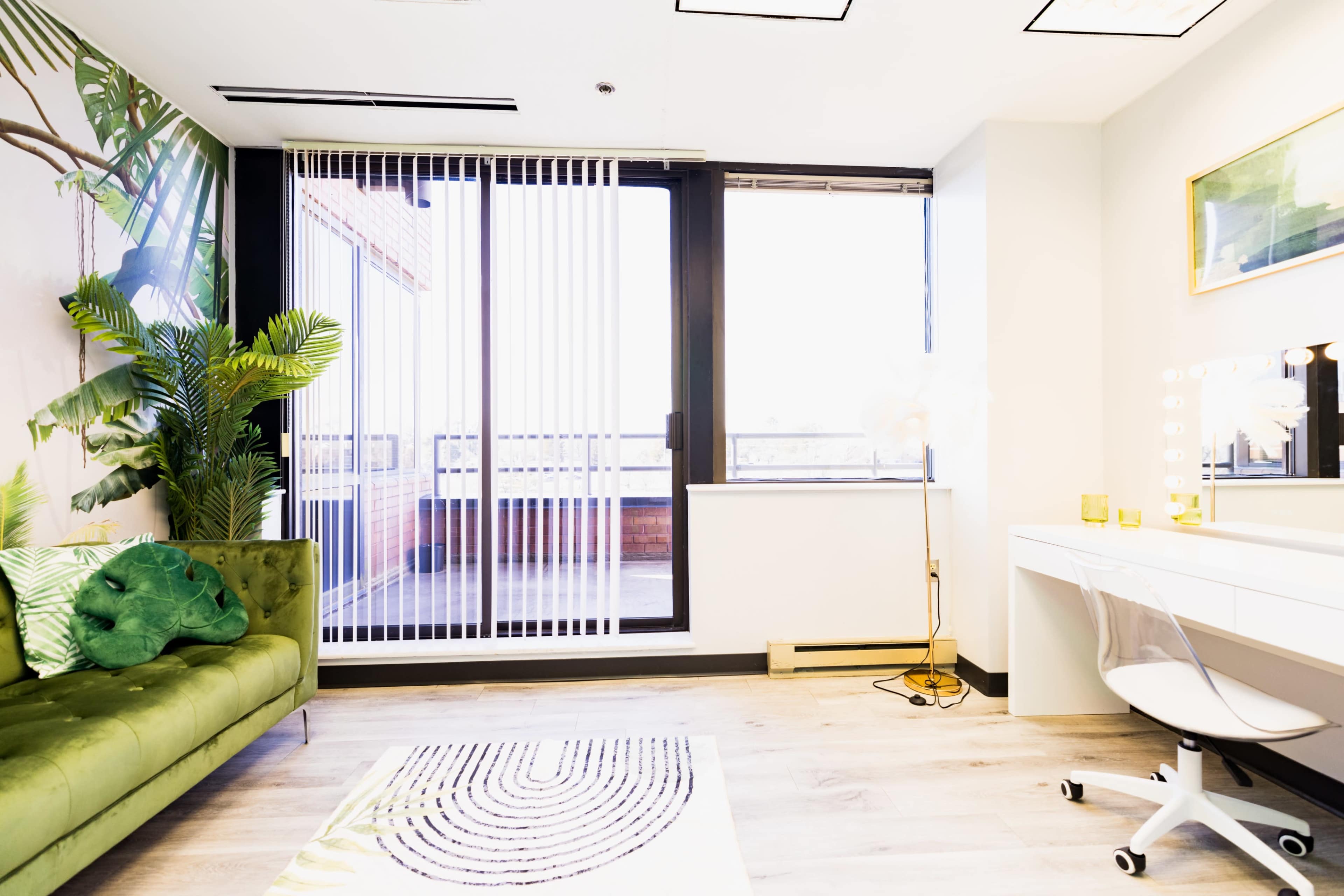 The image shows a bright, modern room with a green velvet sofa, a large window with vertical blinds, and a vanity desk with a circular mirror.
