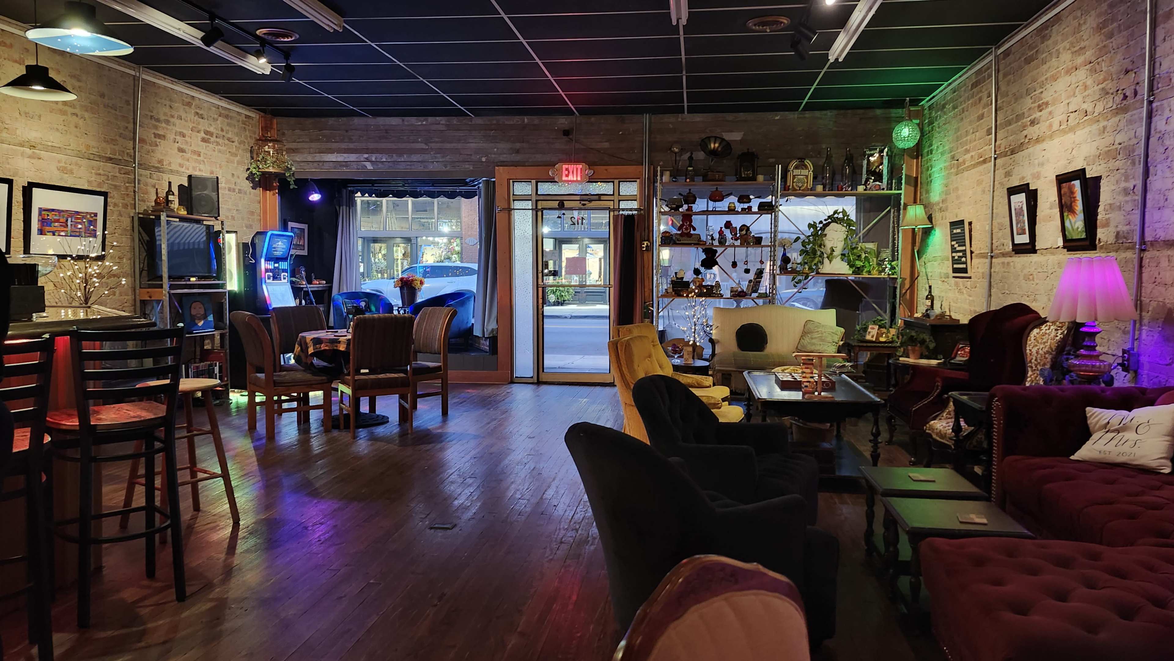 The image shows a cozy cafe interior featuring a mix of seating options, warm lighting, and decor elements, with large windows allowing natural light to enter.