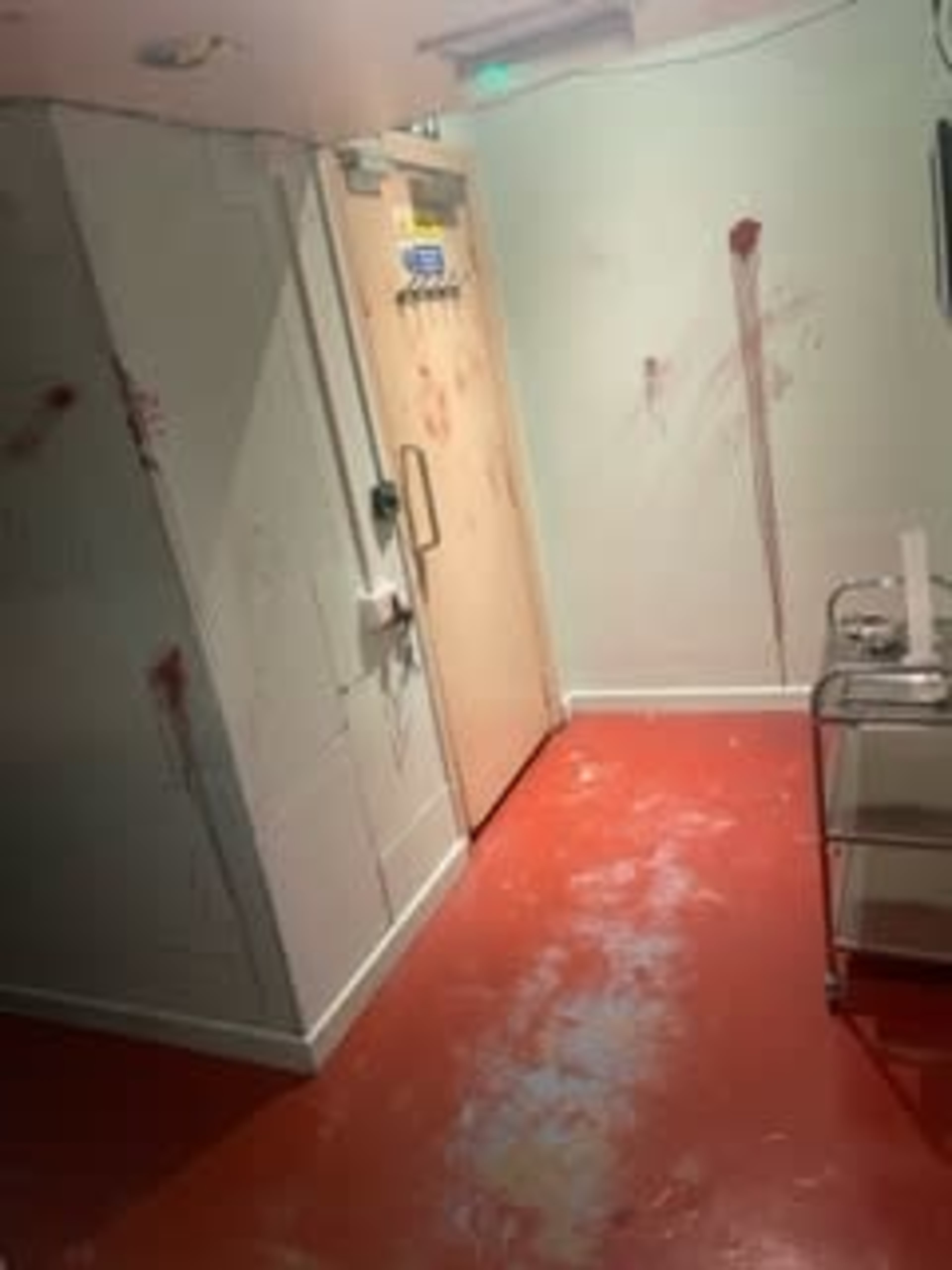 A room with a red floor and walls marked by splatters of what appears to be blood, alongside a closed door and a wheeled cart.