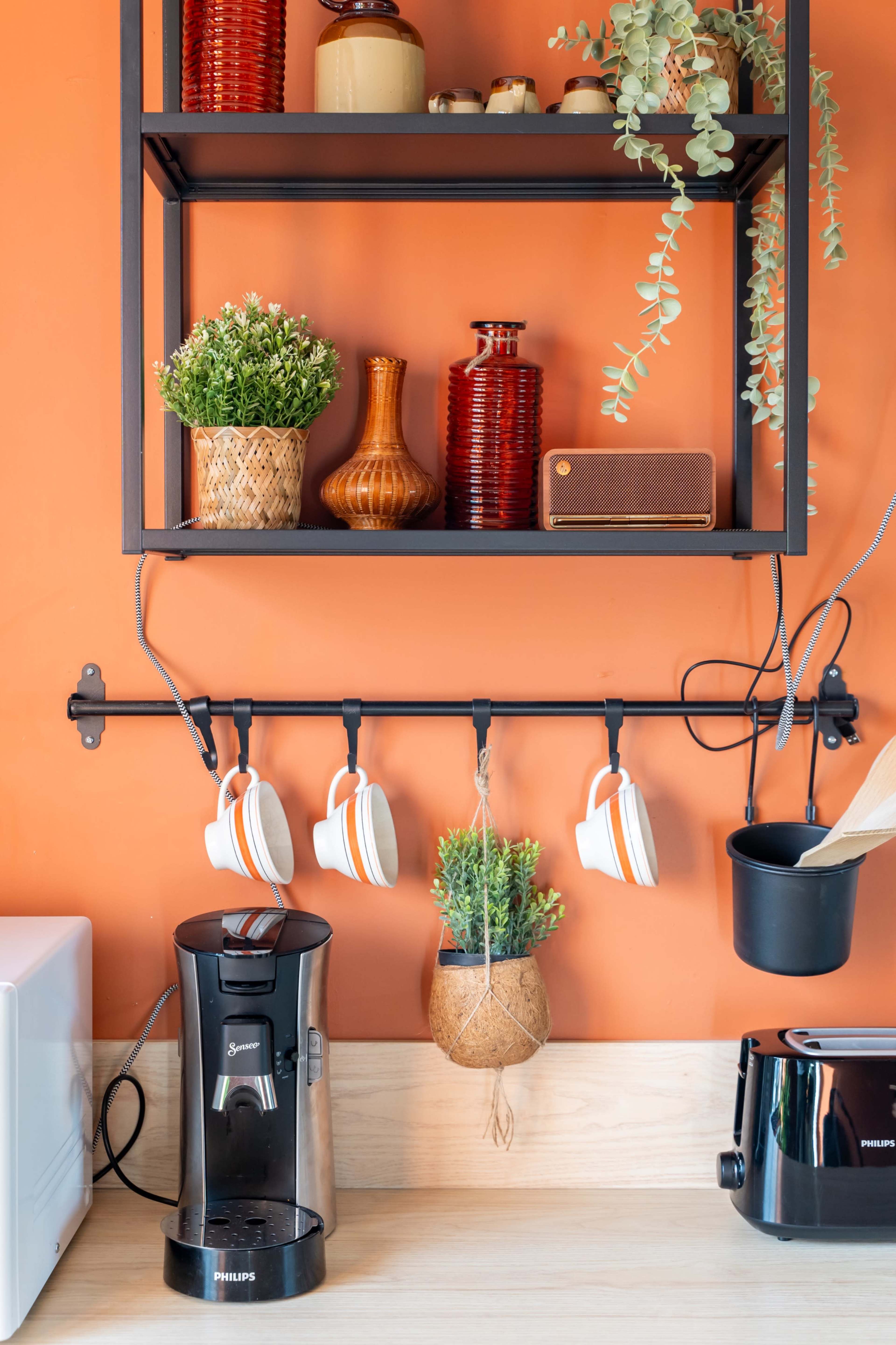 The image shows a kitchen countertop with a coffee maker, a toaster, and a metal rack displaying decorative items and potted plants against an orange wall.