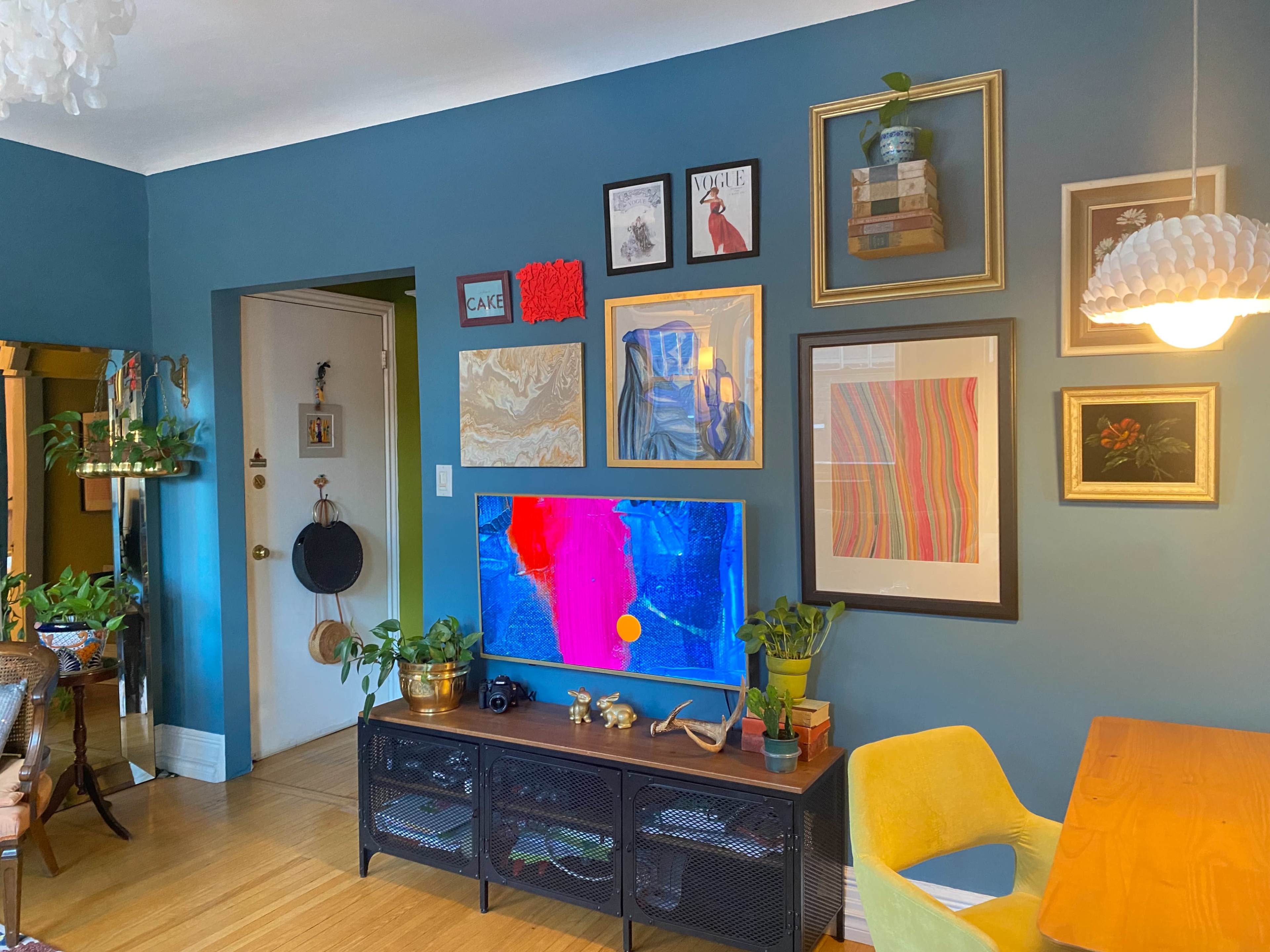 A colorful living room with a blue wall, a television mounted on a wooden stand, and various framed artworks and plants displayed on the walls and furniture.