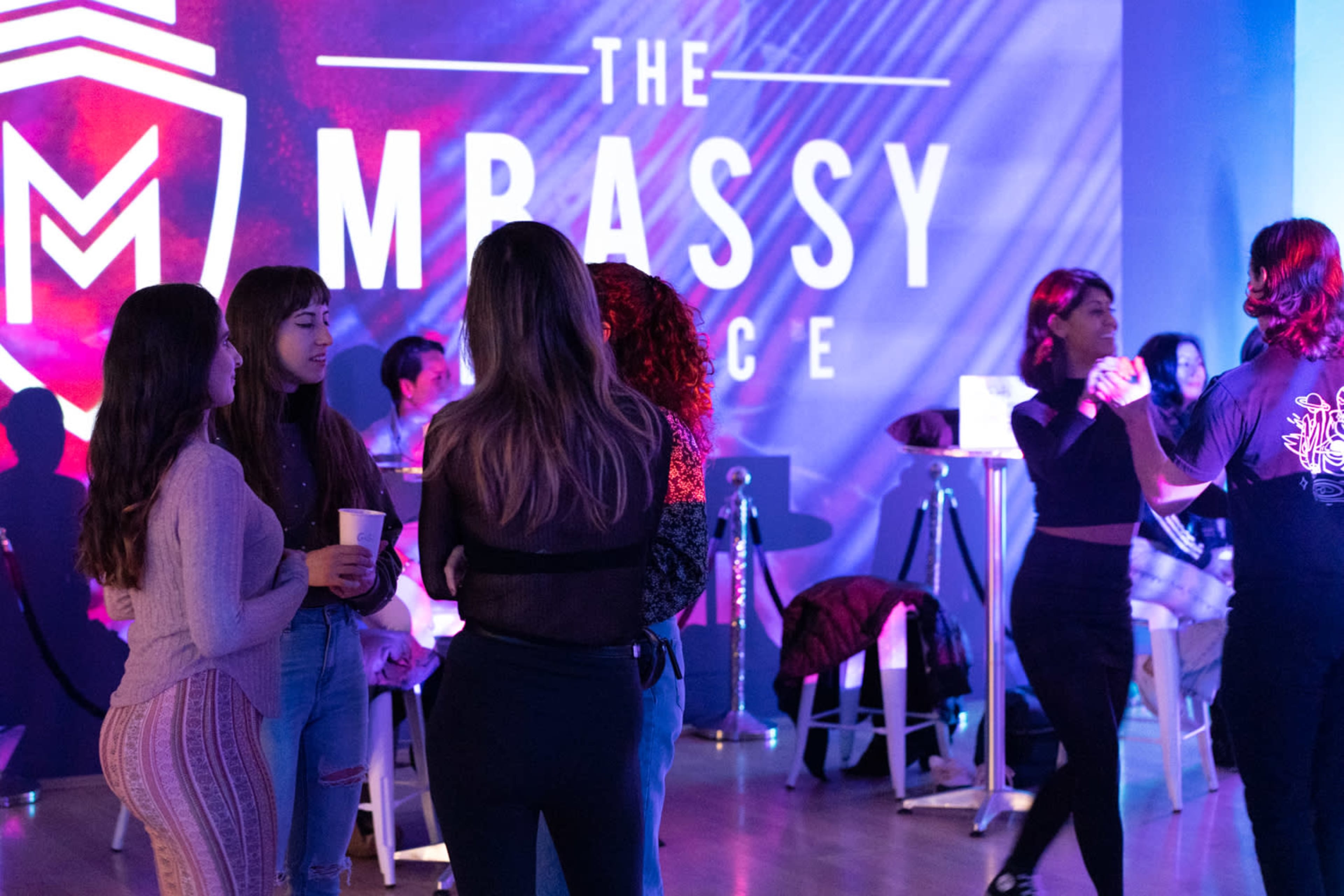 A group of people socializes in front of a large sign that reads "THE MRASSY PLACE," with some individuals dancing in the background.