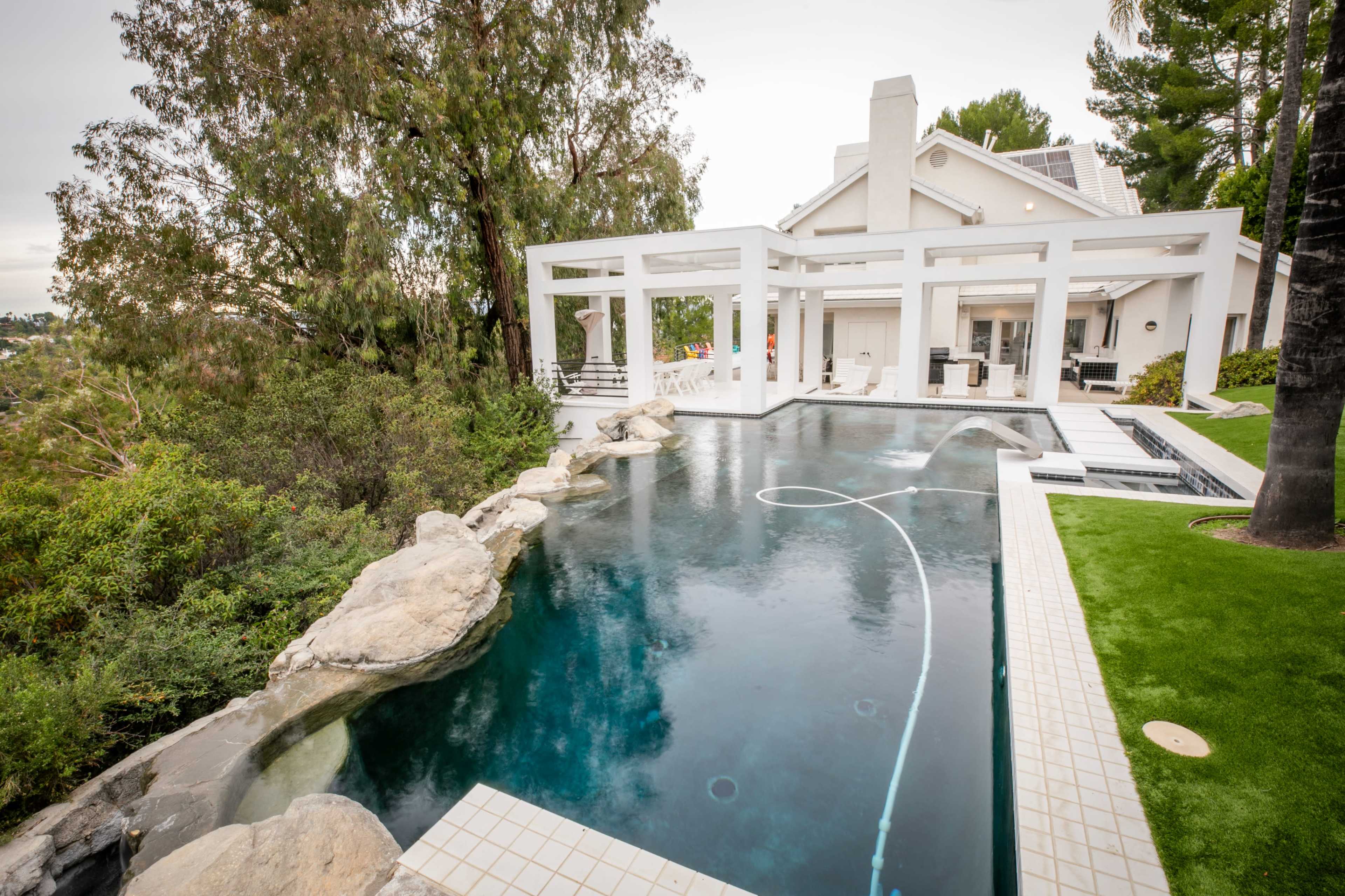 The image shows a modern house with a large swimming pool, surrounded by greenery and featuring a rocky landscape and outdoor seating area.
