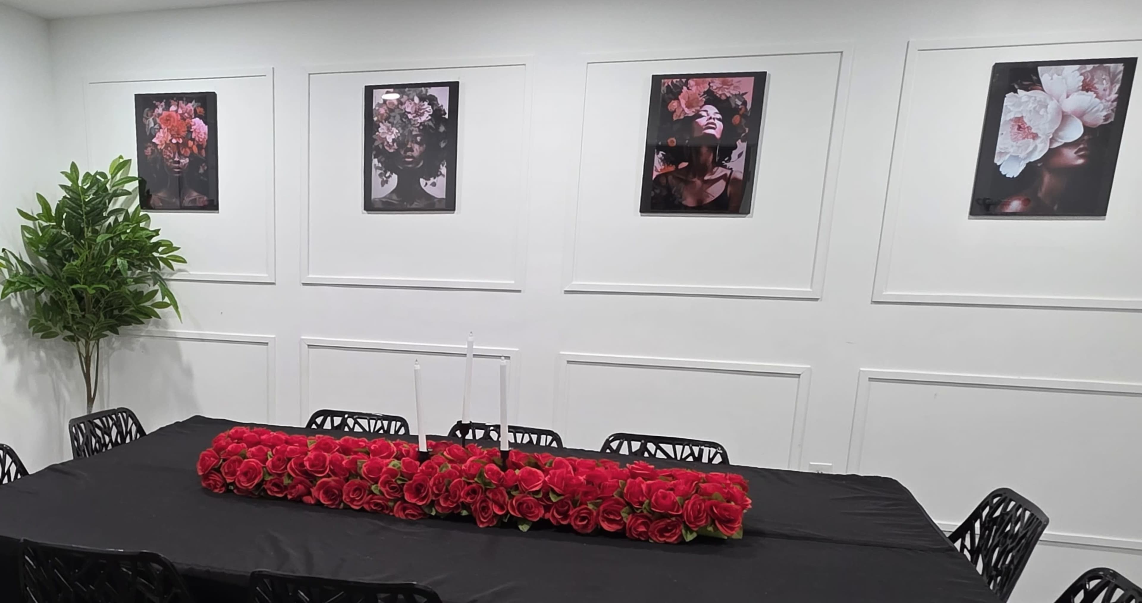 A long table adorned with a red rose centerpiece and white candles is positioned in a room featuring framed floral portraits on the walls.