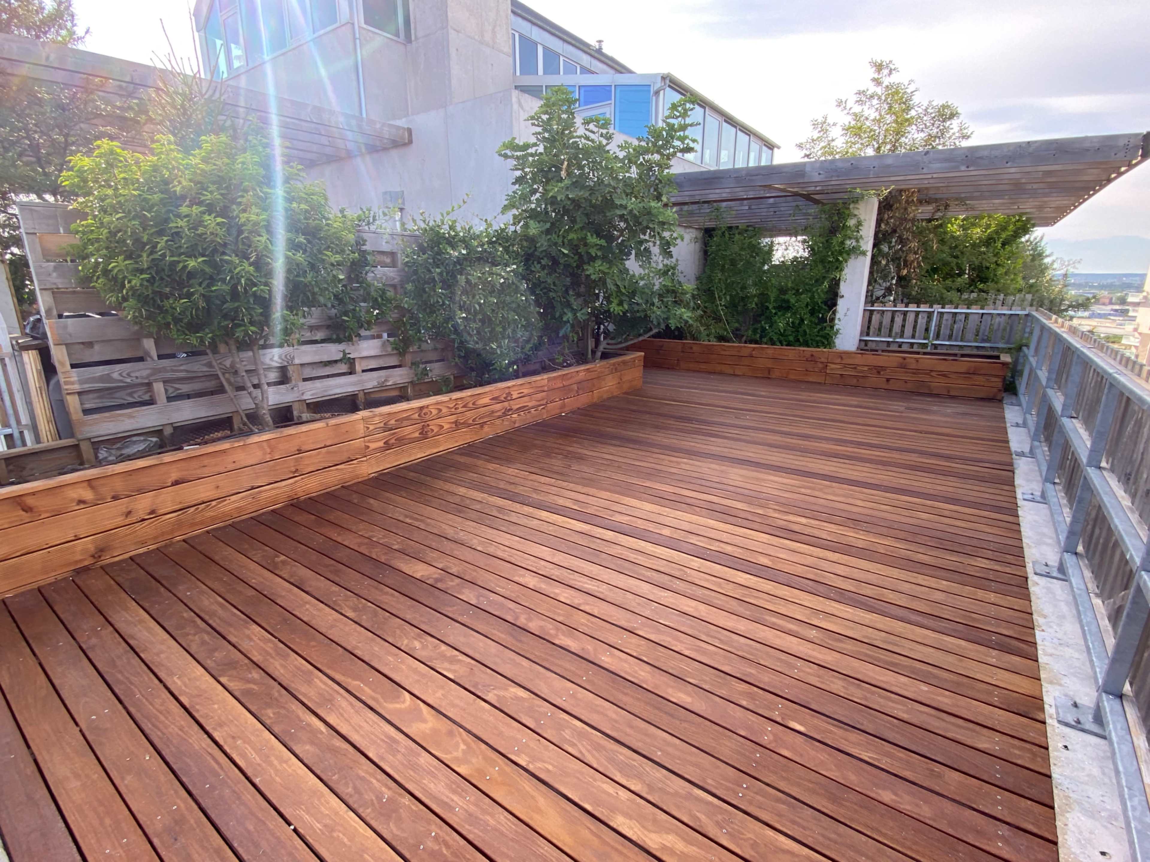 The image shows a spacious wooden deck surrounded by greenery and a pergola.