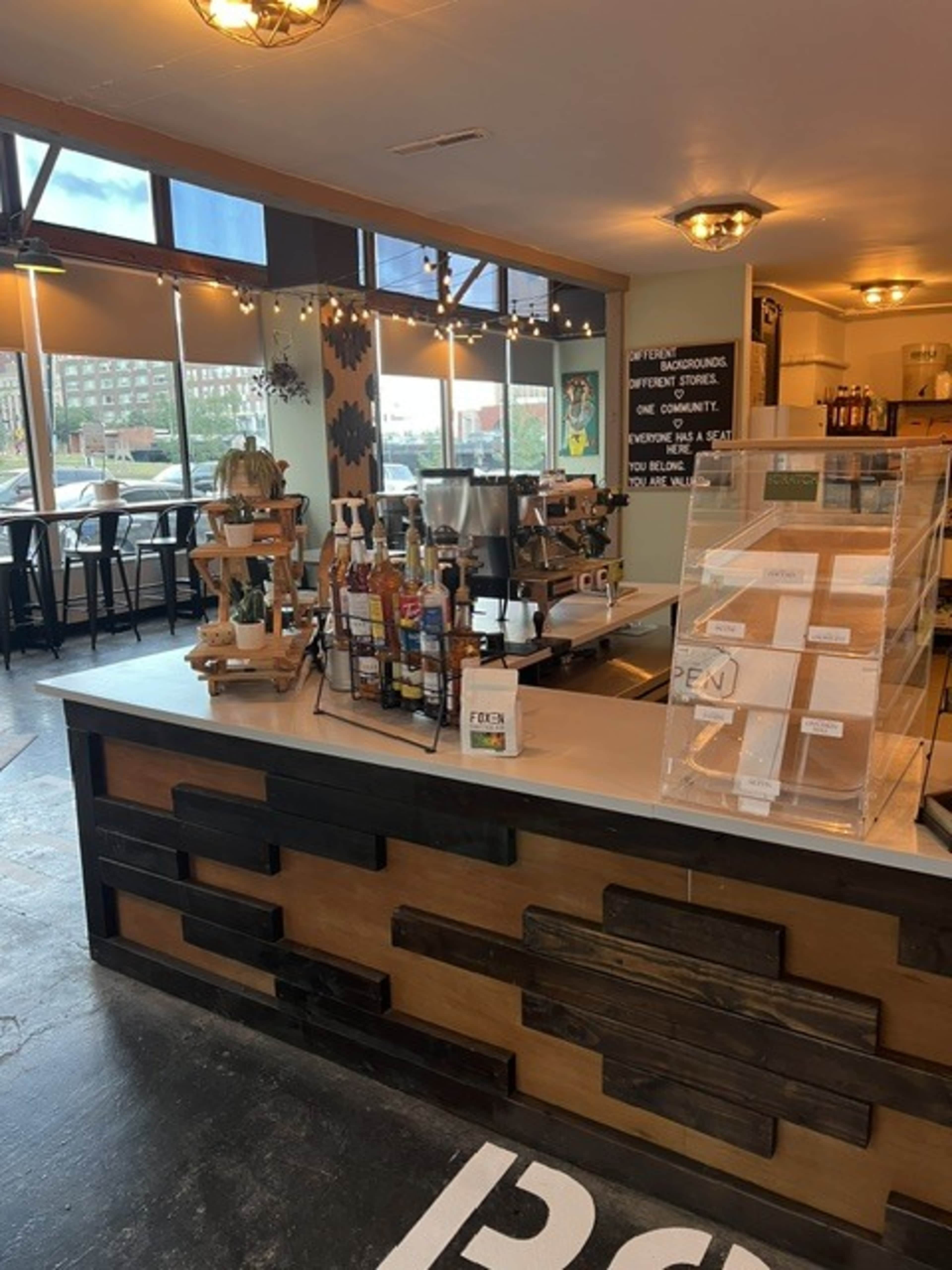 The image shows the interior of a café featuring a service counter with various drink dispensers, seating in the background, and large windows allowing natural light to enter.