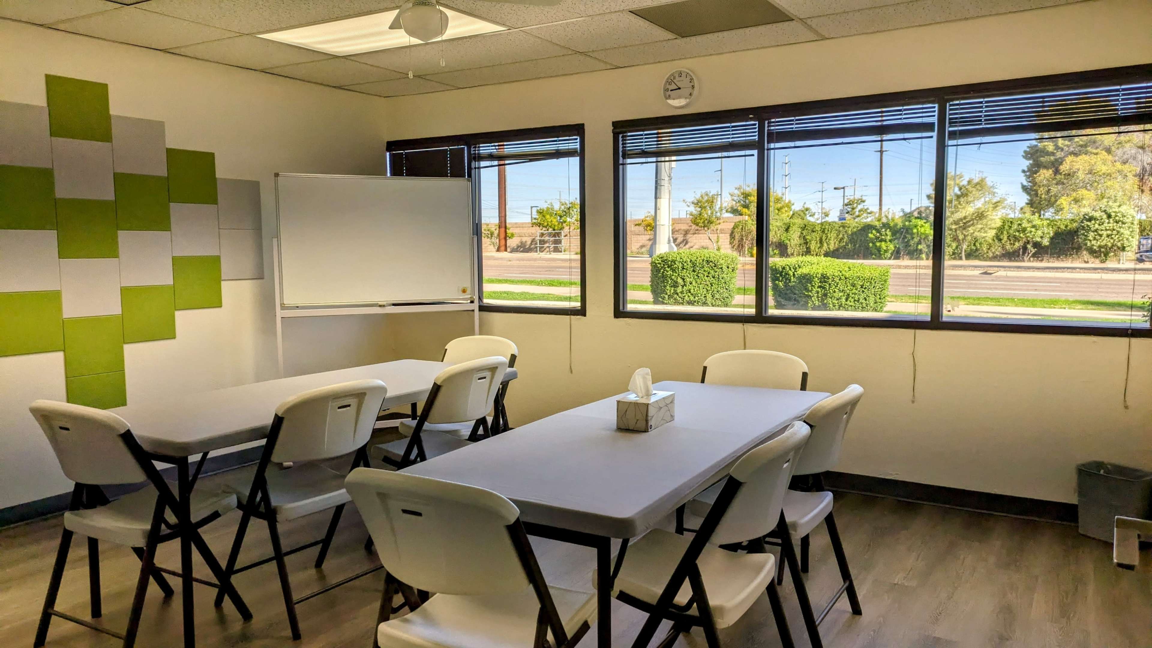 A bright classroom with several folding tables and chairs arranged, a whiteboard, and large windows letting in natural light.