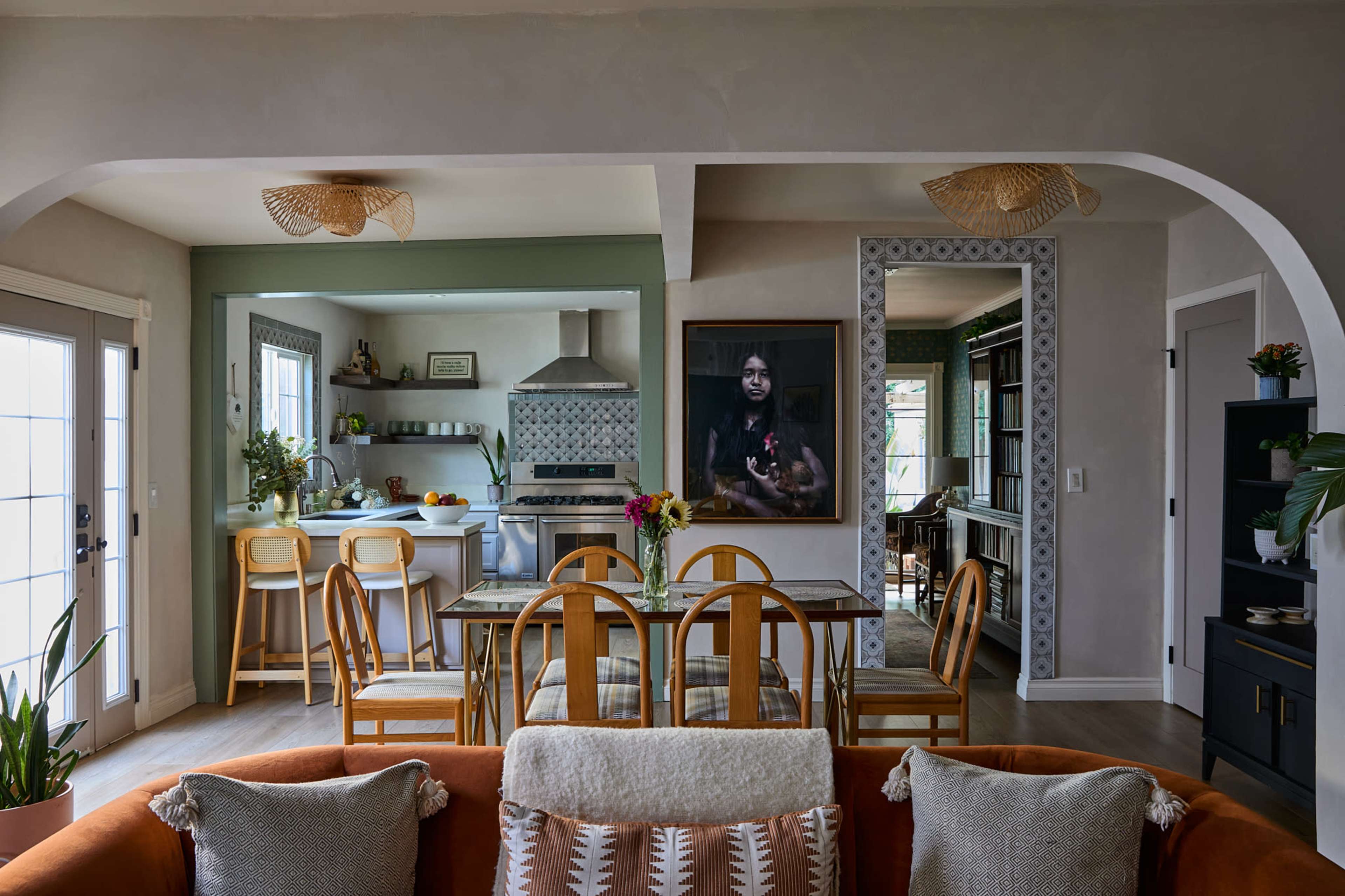 The image shows a modern living space featuring a kitchen visible through an archway, with a dining table, stylish decor, and a large portrait on the wall.