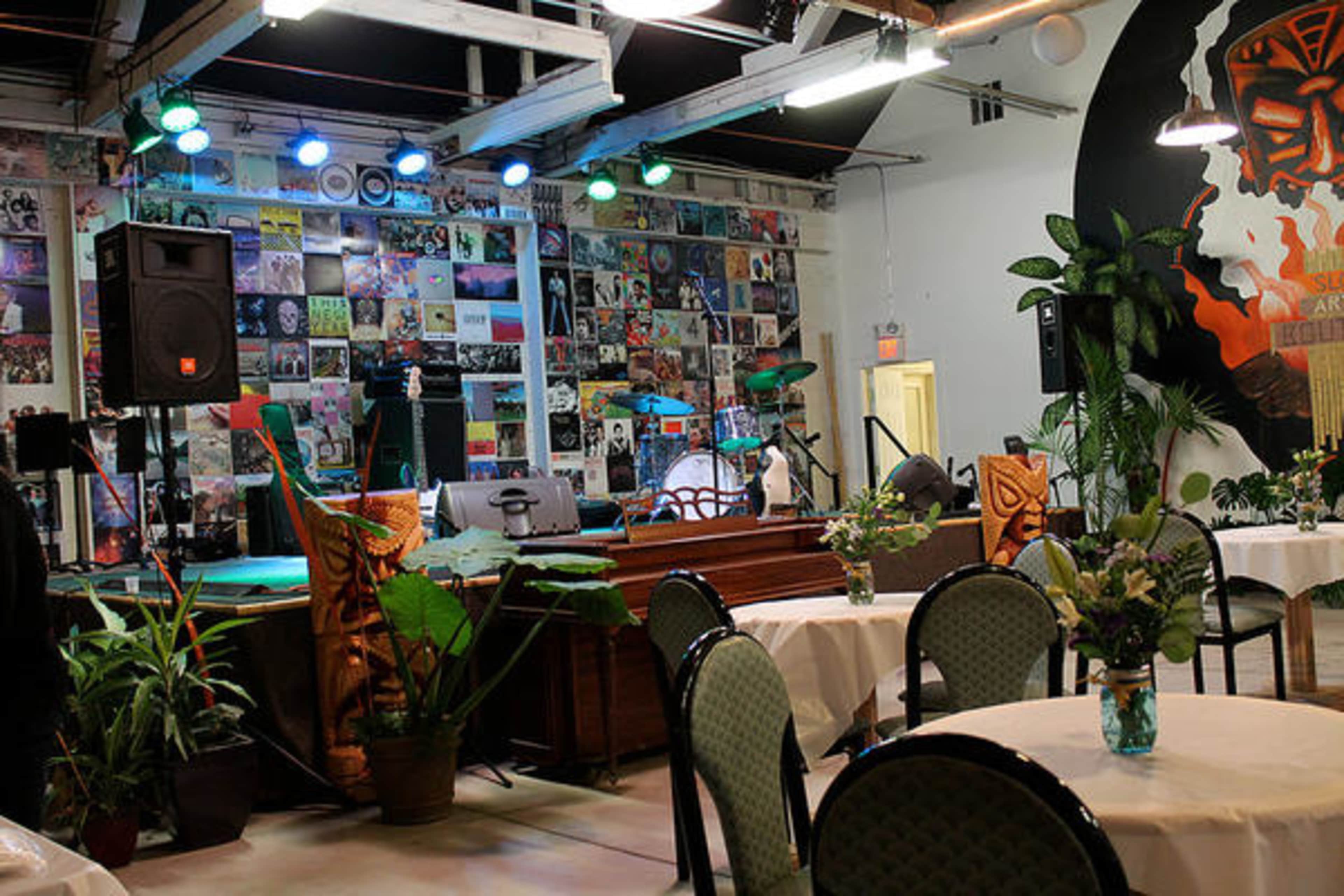 A music venue with a stage set up for a performance, surrounded by plants and tables arranged for patrons.