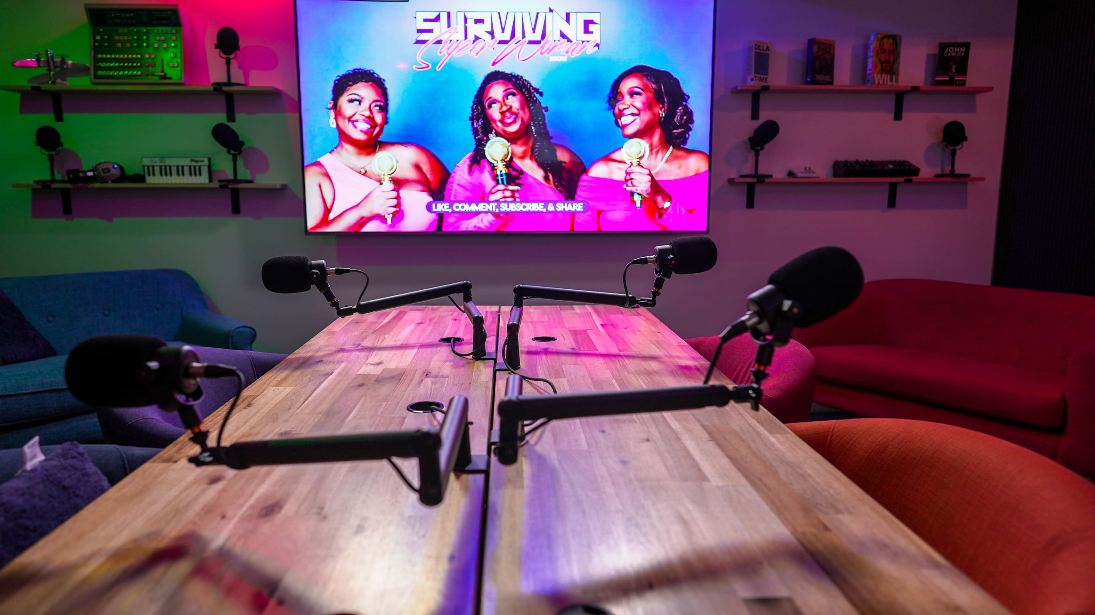 A recording studio setup with a wooden table, two microphones on each side, and a large screen displaying the title "Surviving Super Chicas."
