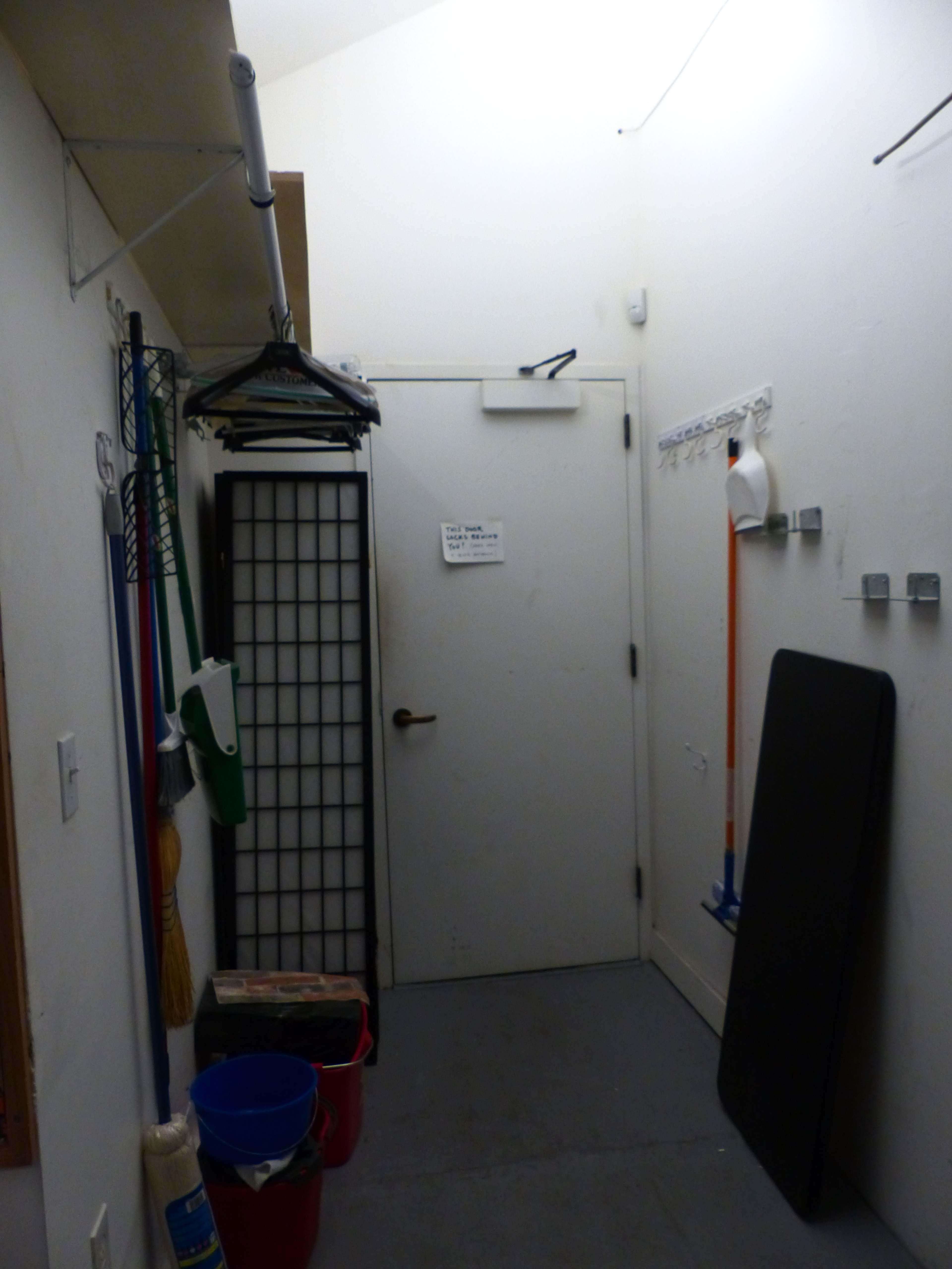 A narrow hallway features a closed door, cleaning tools hung on the wall, and a couple of storage containers on the floor.