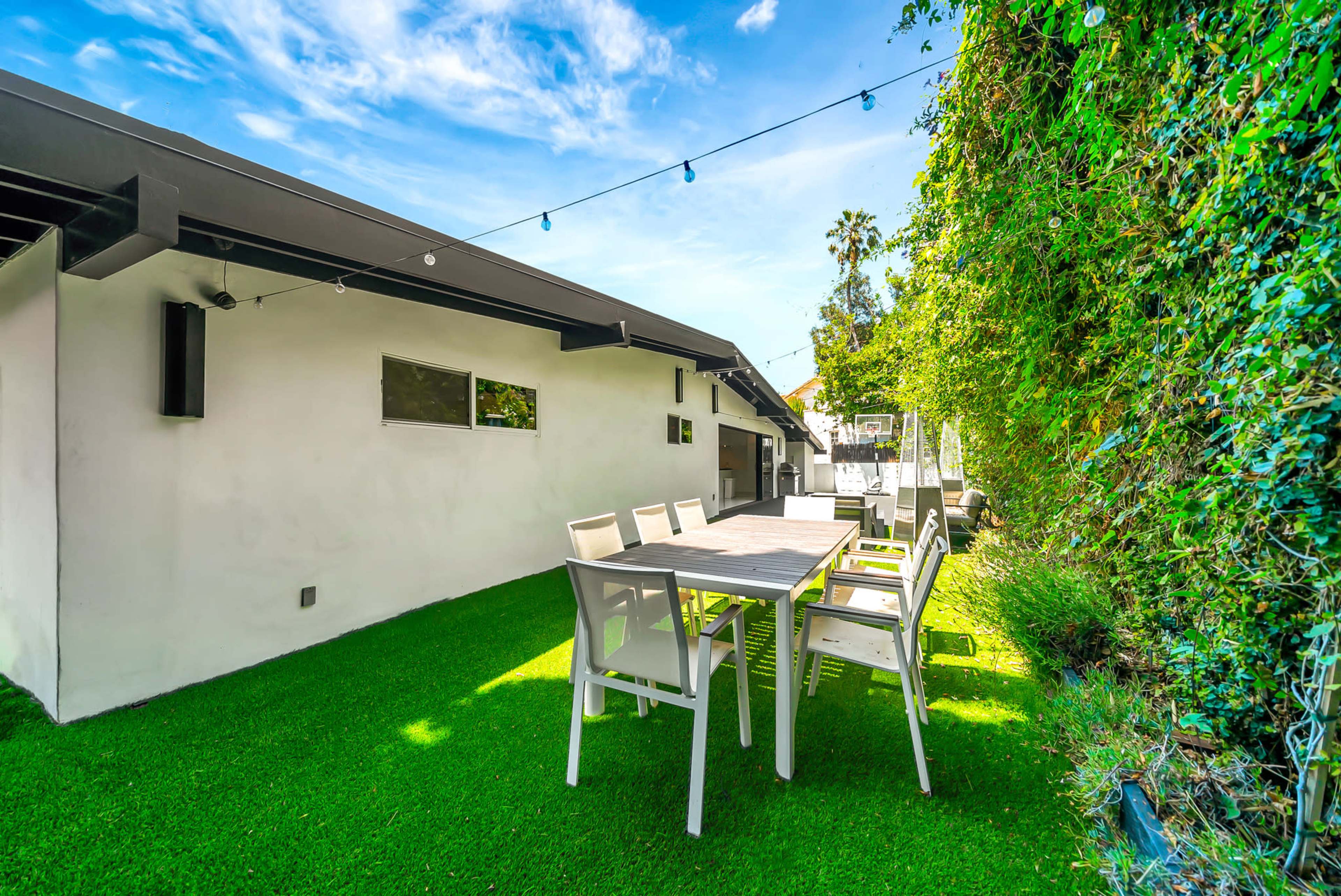 A dining area with a long table and chairs is set on lush green grass beside a modern white house, with string lights overhead and greenery along the wall.