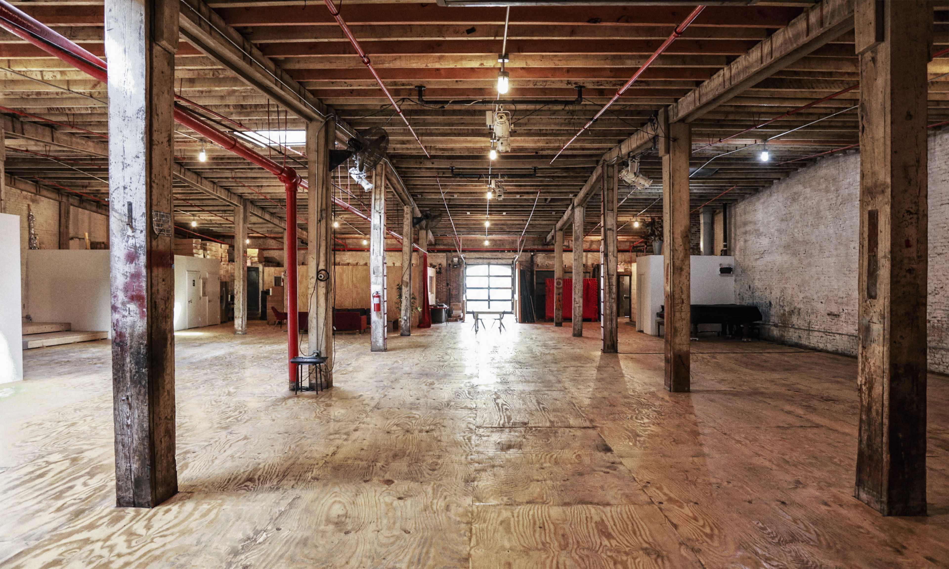 The image shows an expansive, empty industrial space with wooden floors and exposed beams, featuring a row of columns and a large rear window letting in light.