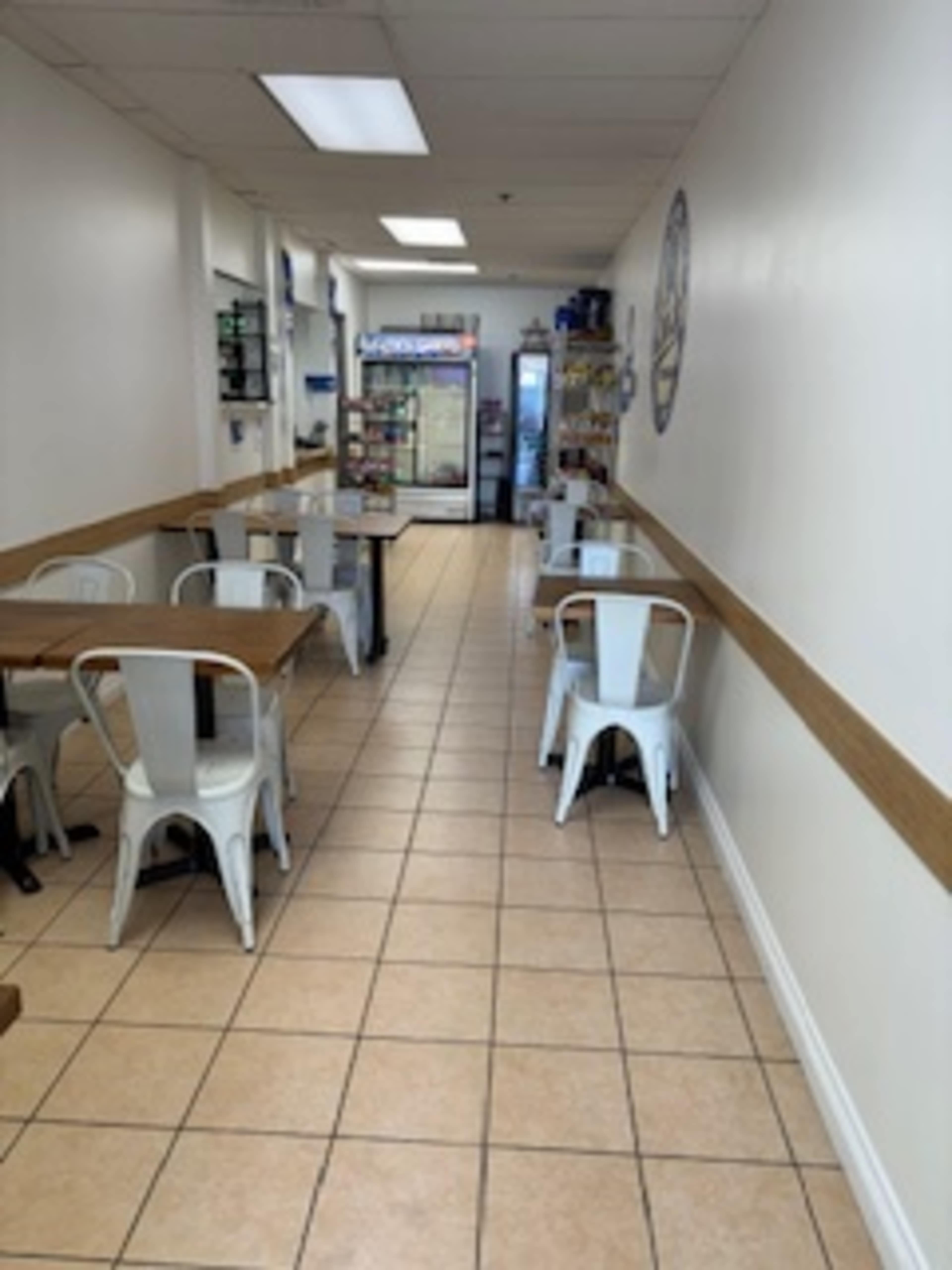 The image shows a narrow dining area with metal tables and chairs, lined with tiled flooring, and leading to a cooler at the far end.