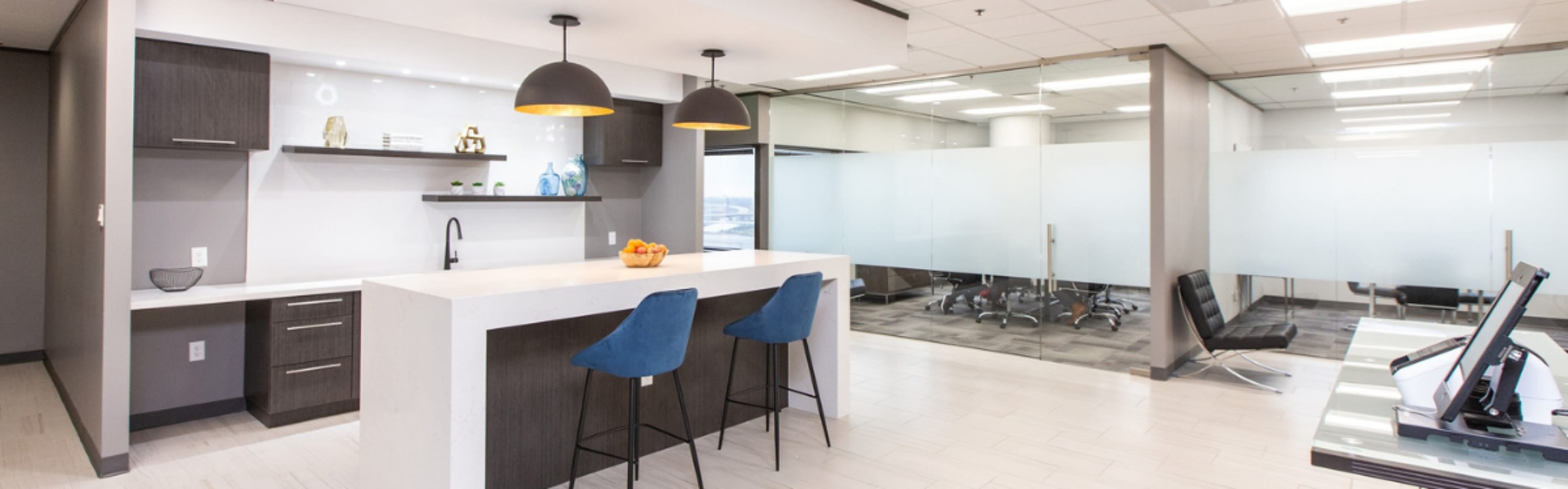The image shows a modern office space with a kitchen area featuring a bar counter, stools, and a glass-walled conference room in the background.