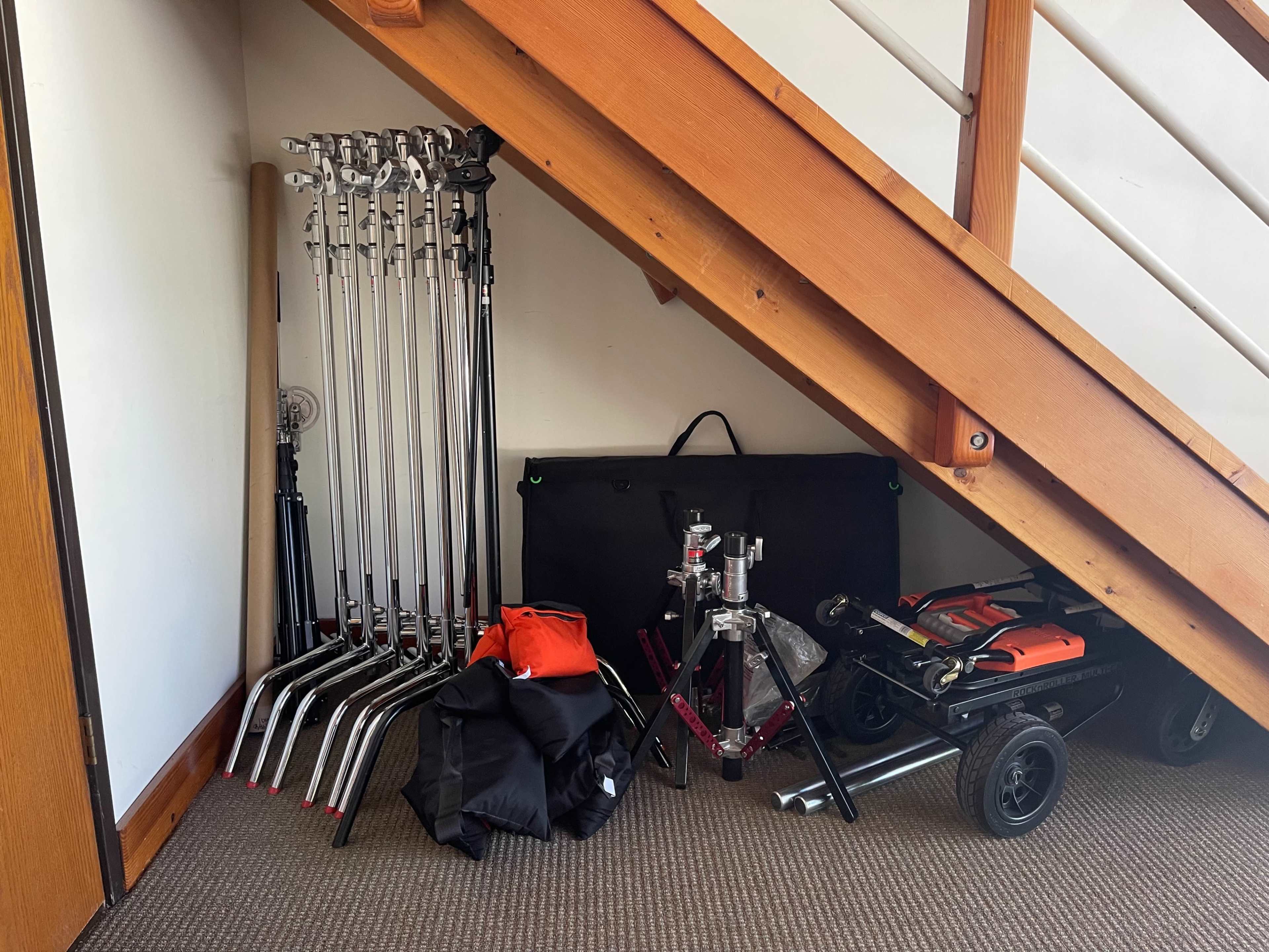 A storage area under a staircase containing various photography equipment, including light stands, a black backdrop, and a wheeled cart.