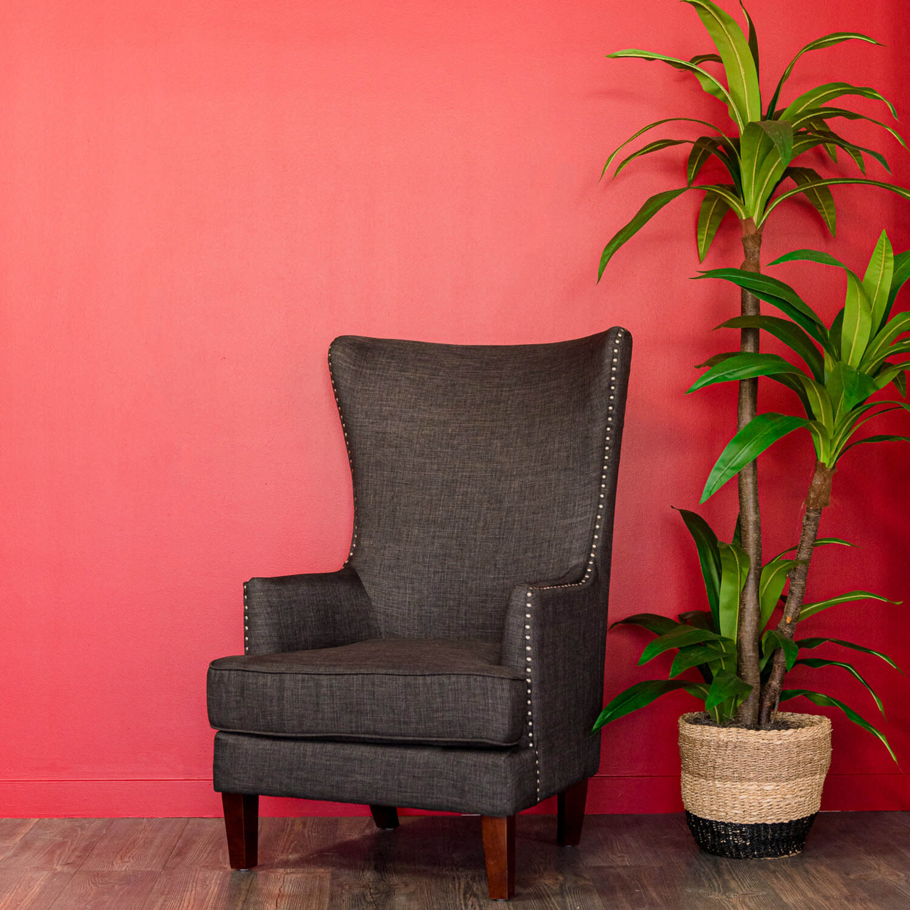 A gray wingback chair with silver nailhead trim is positioned next to a potted plant against a red wall.