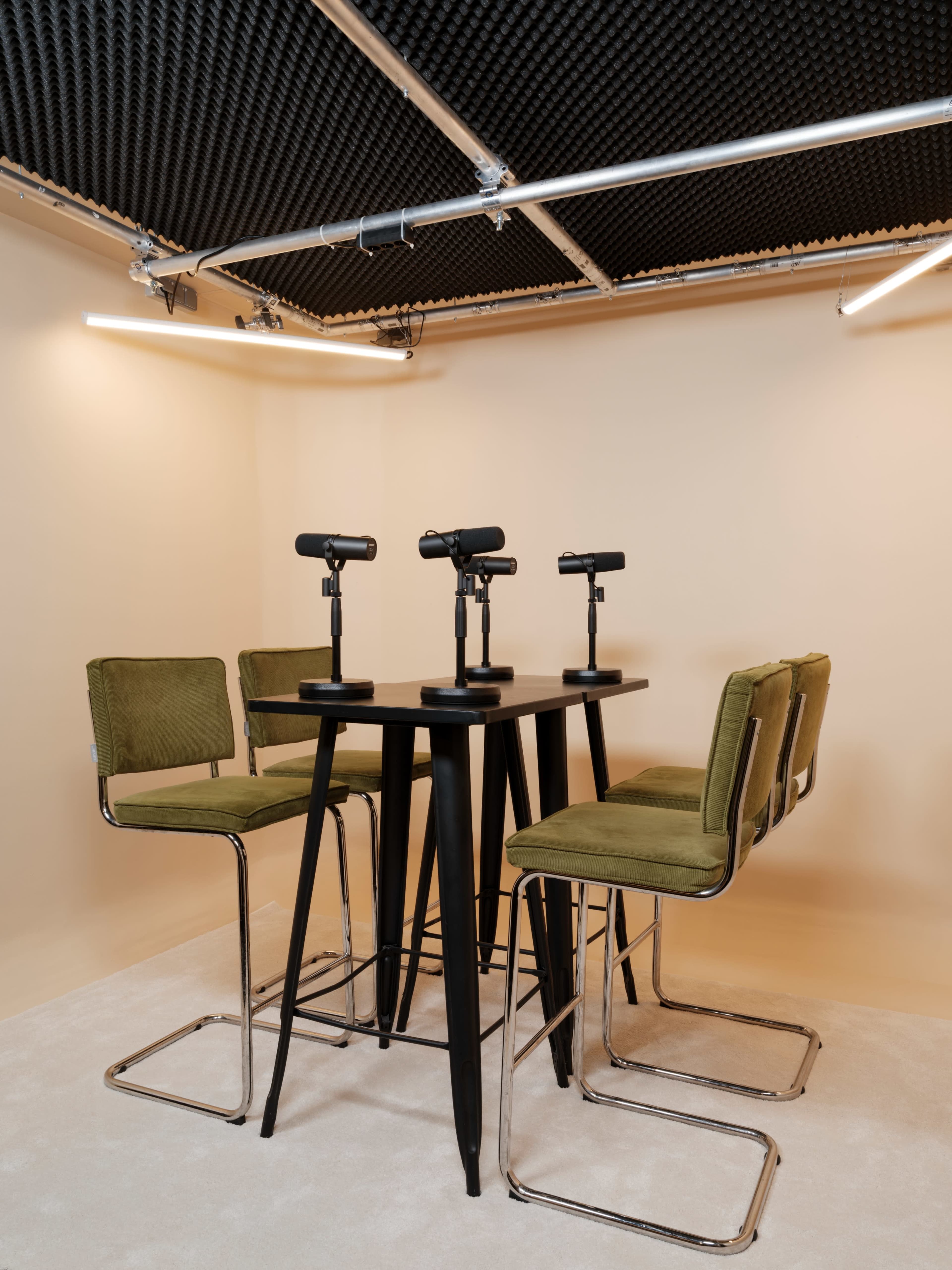 A set of six green barstools with microphones is arranged around a black table in a soundproofed room.
