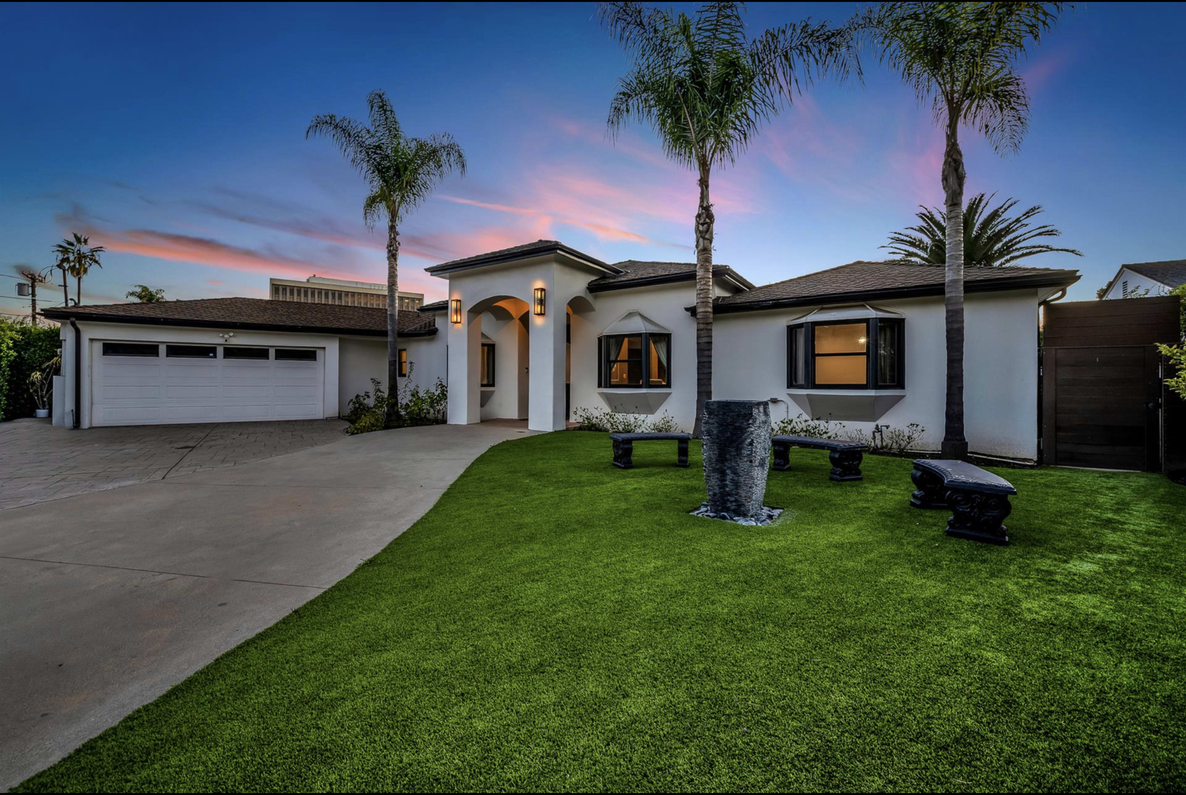 The image shows a modern single-story house with a manicured green lawn, palm trees, and a concrete driveway leading to a three-car garage.