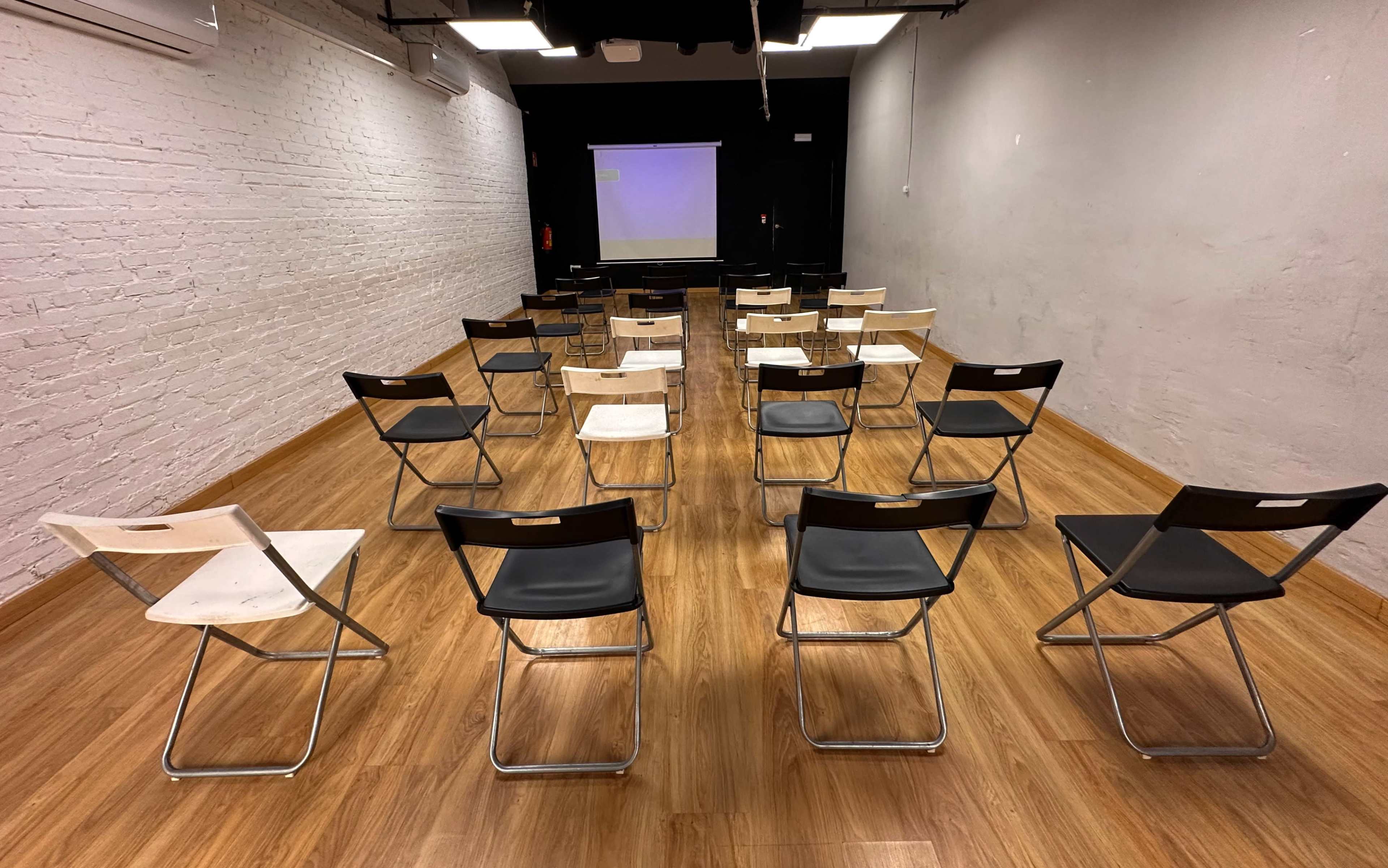 The image shows a room set up for a presentation, featuring rows of folding chairs facing a blank screen at the front.