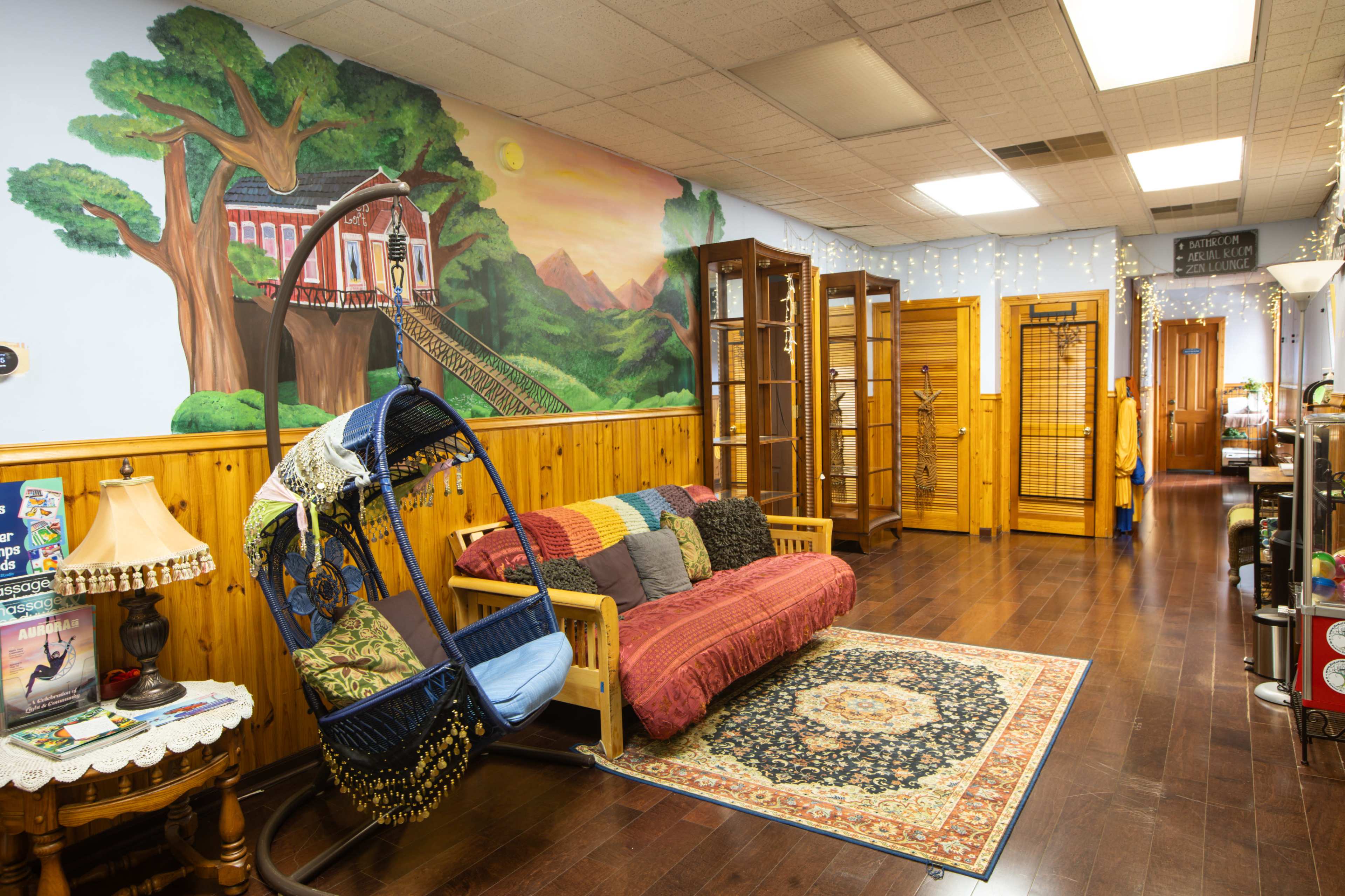 The image shows a warmly decorated room with a mural of a landscape on one wall, a swing chair, a vibrant rug in the center, and wooden furniture along the walls.