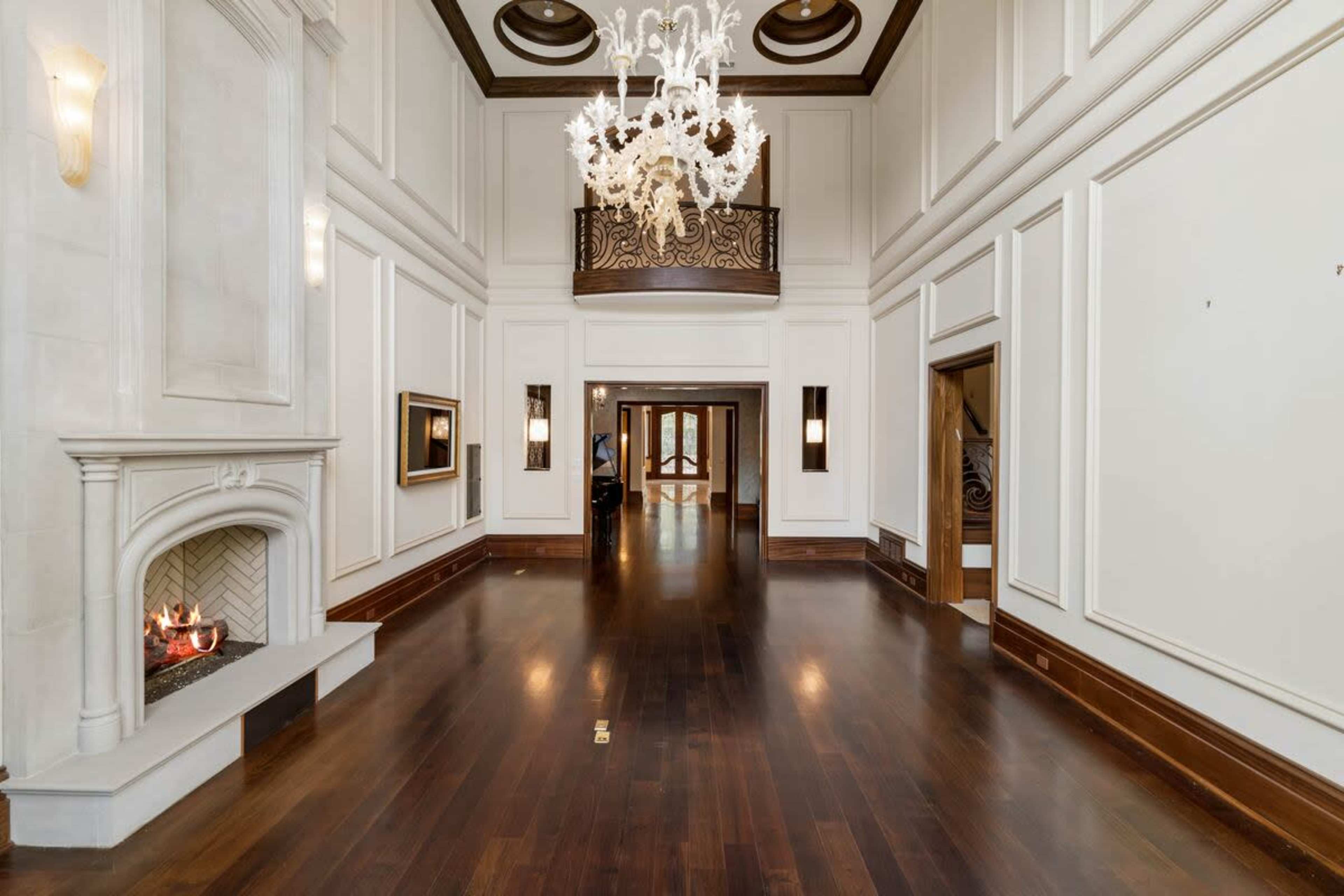The image shows a spacious, elegantly designed interior of a house featuring a chandelier, fireplace, and wooden flooring.