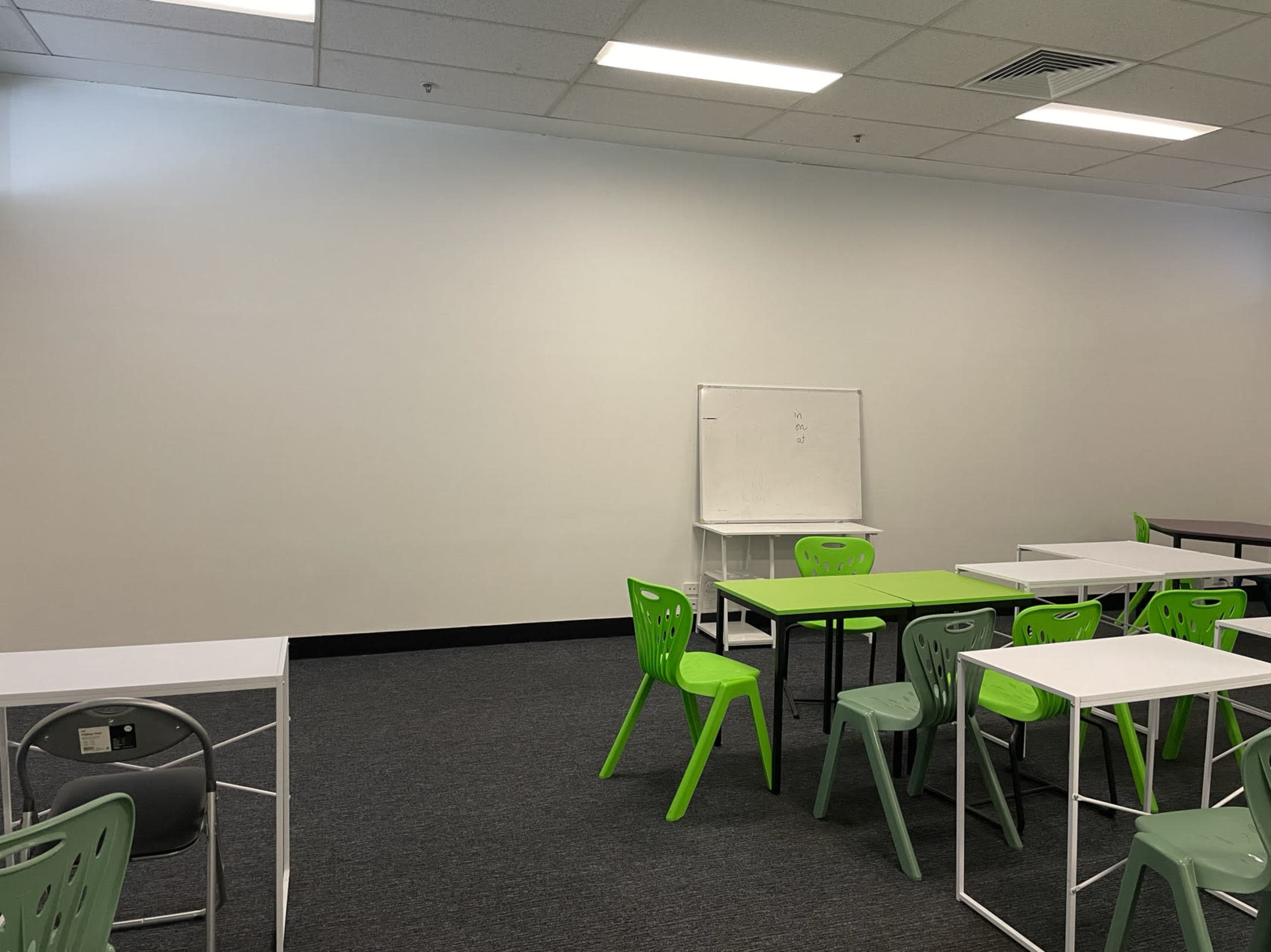An empty classroom features green chairs and tables arranged against a plain wall with a whiteboard.