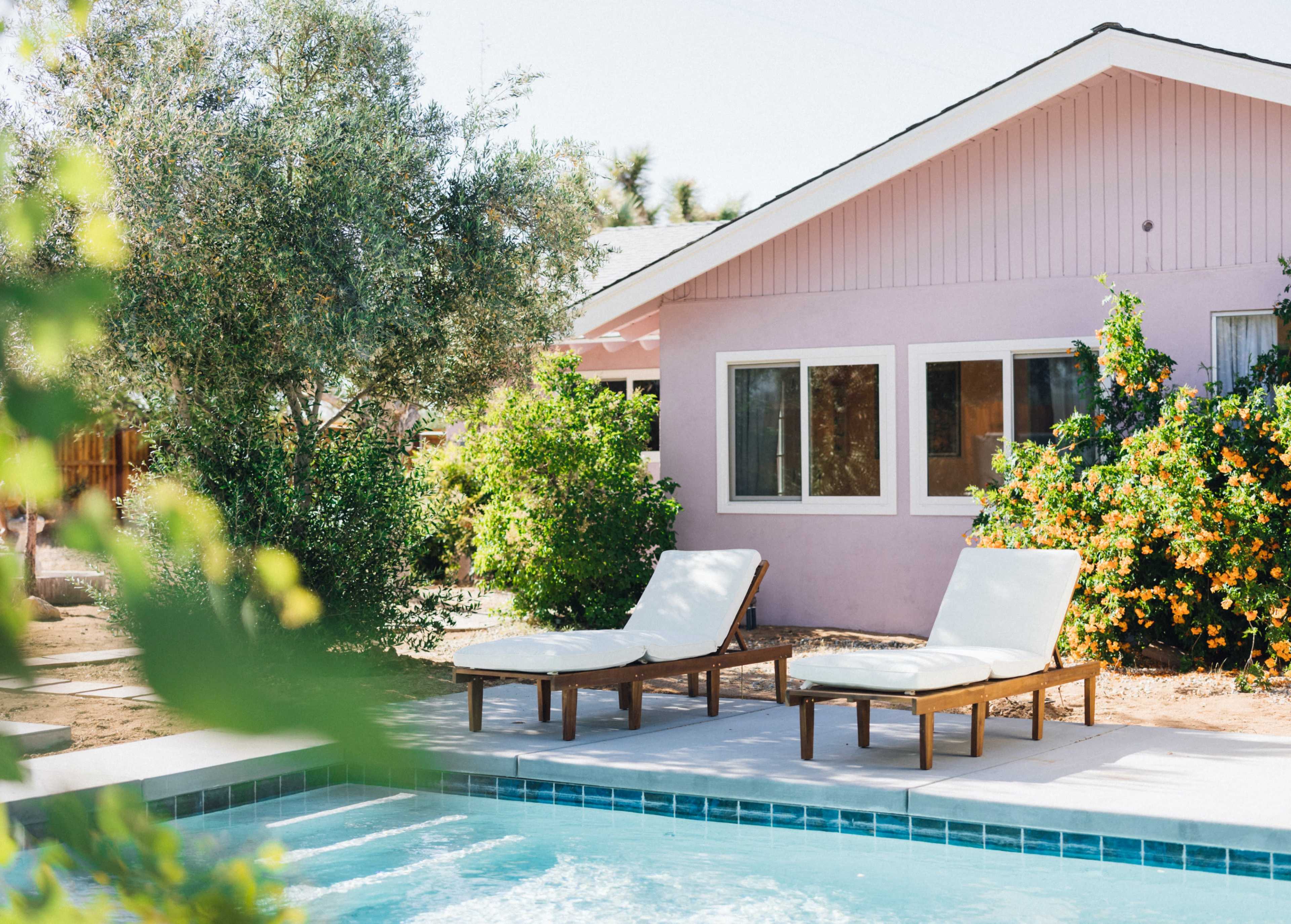 Two white loungers sit beside a clear swimming pool in front of a pink house surrounded by greenery.