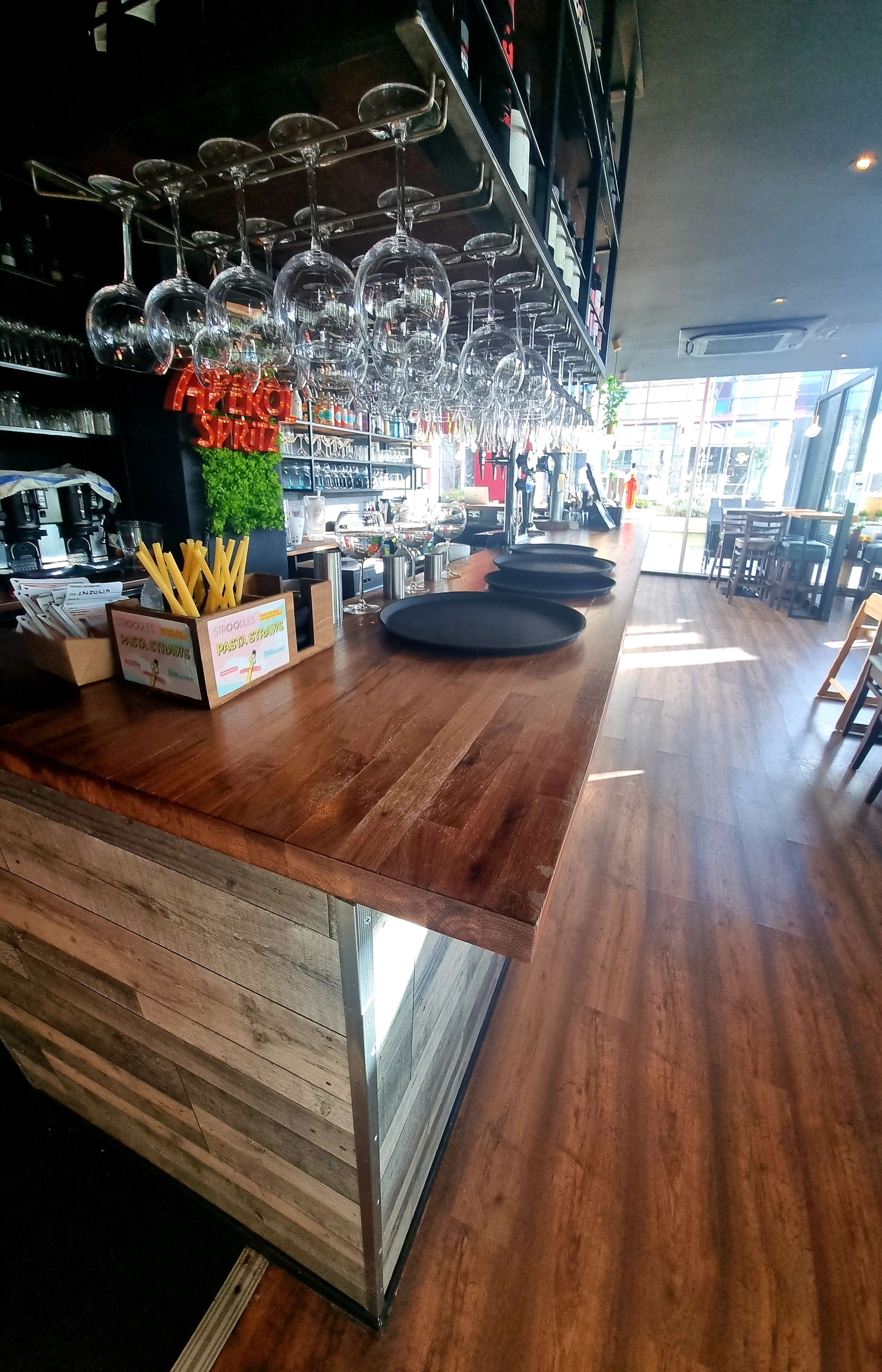 A wooden bar counter with several shiny wine glasses hanging upside down above it and a row of stools in a brightly lit space.