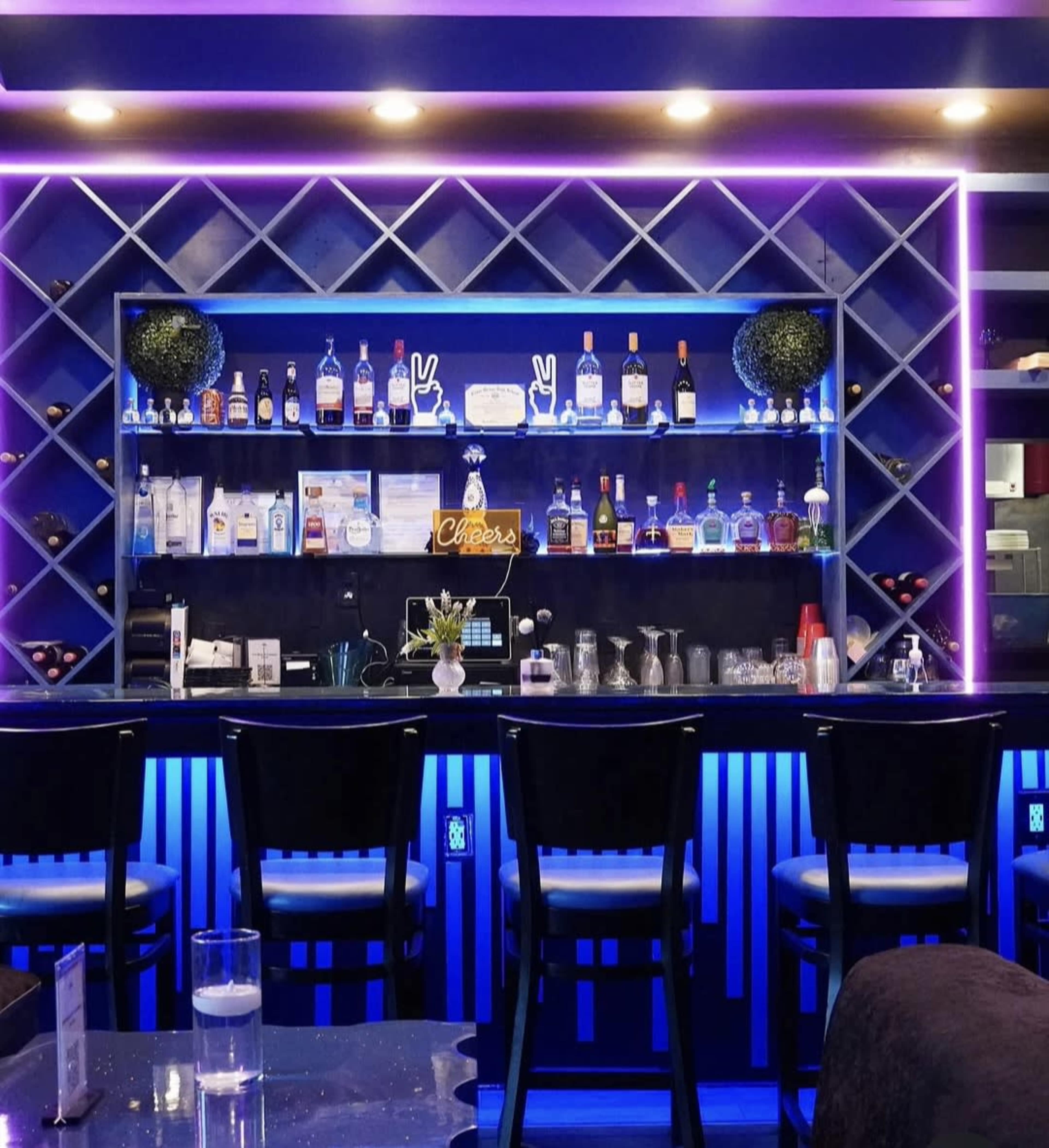 A brightly lit bar displays an array of liquor bottles on a shelf with purple neon accents and black seating in the foreground.