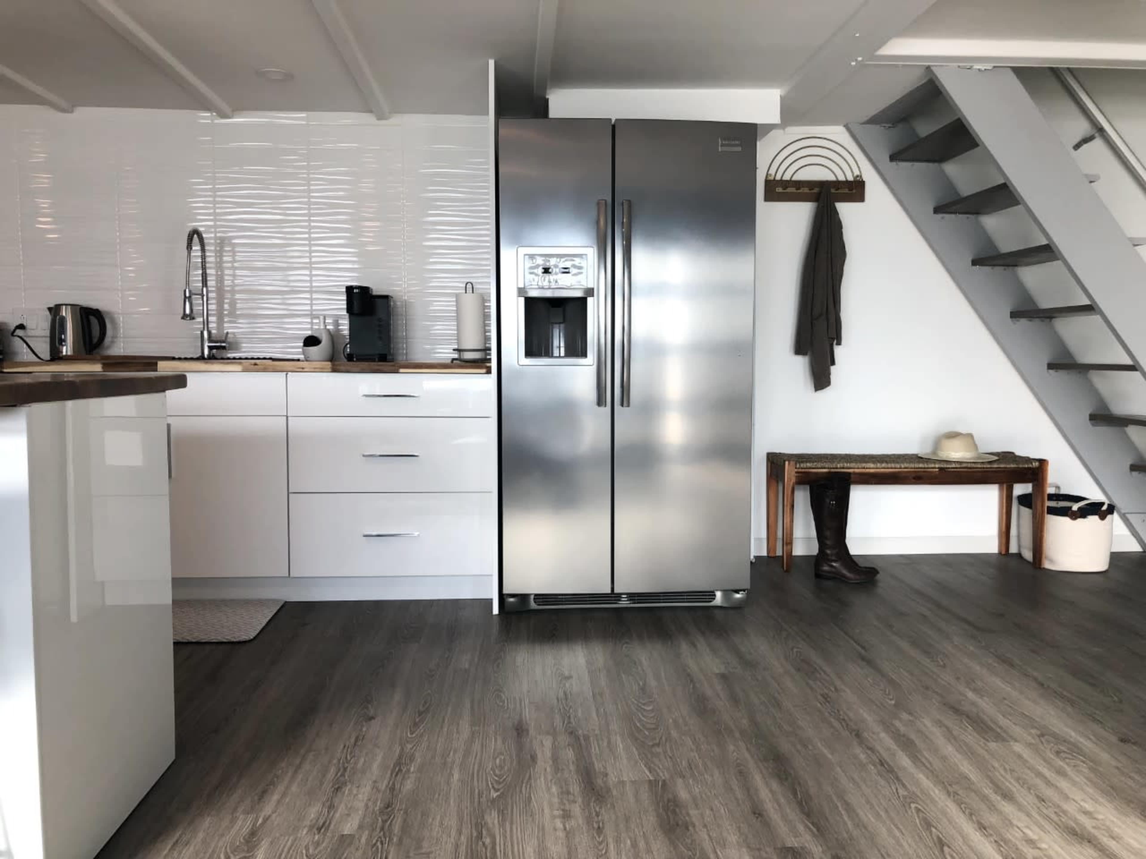 A modern kitchen area with a stainless steel refrigerator, wooden counter, and a staircase in a well-lit room.