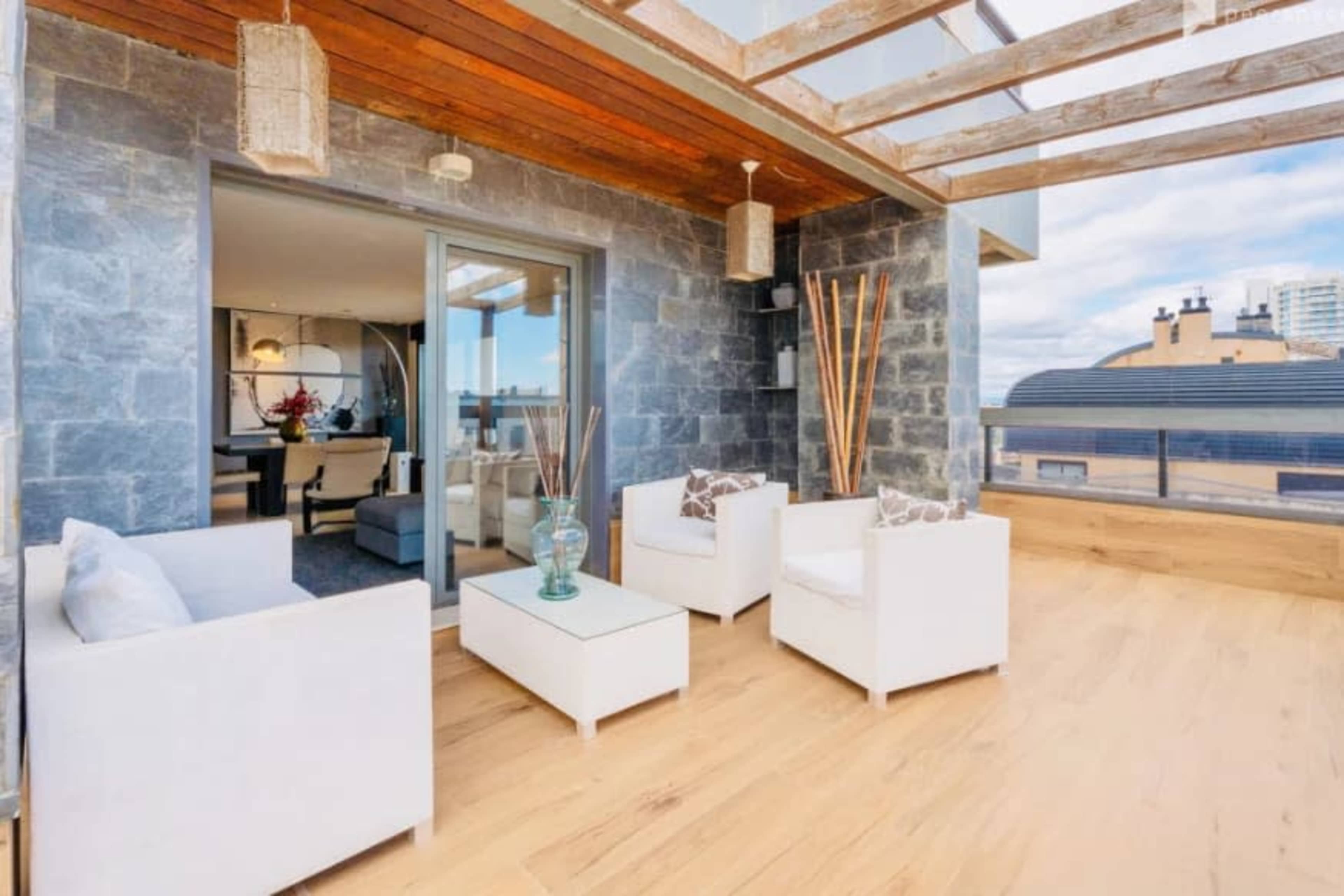 The image shows a modern balcony featuring white outdoor furniture, a glass table, and a stone wall backdrop with a view of a city skyline.