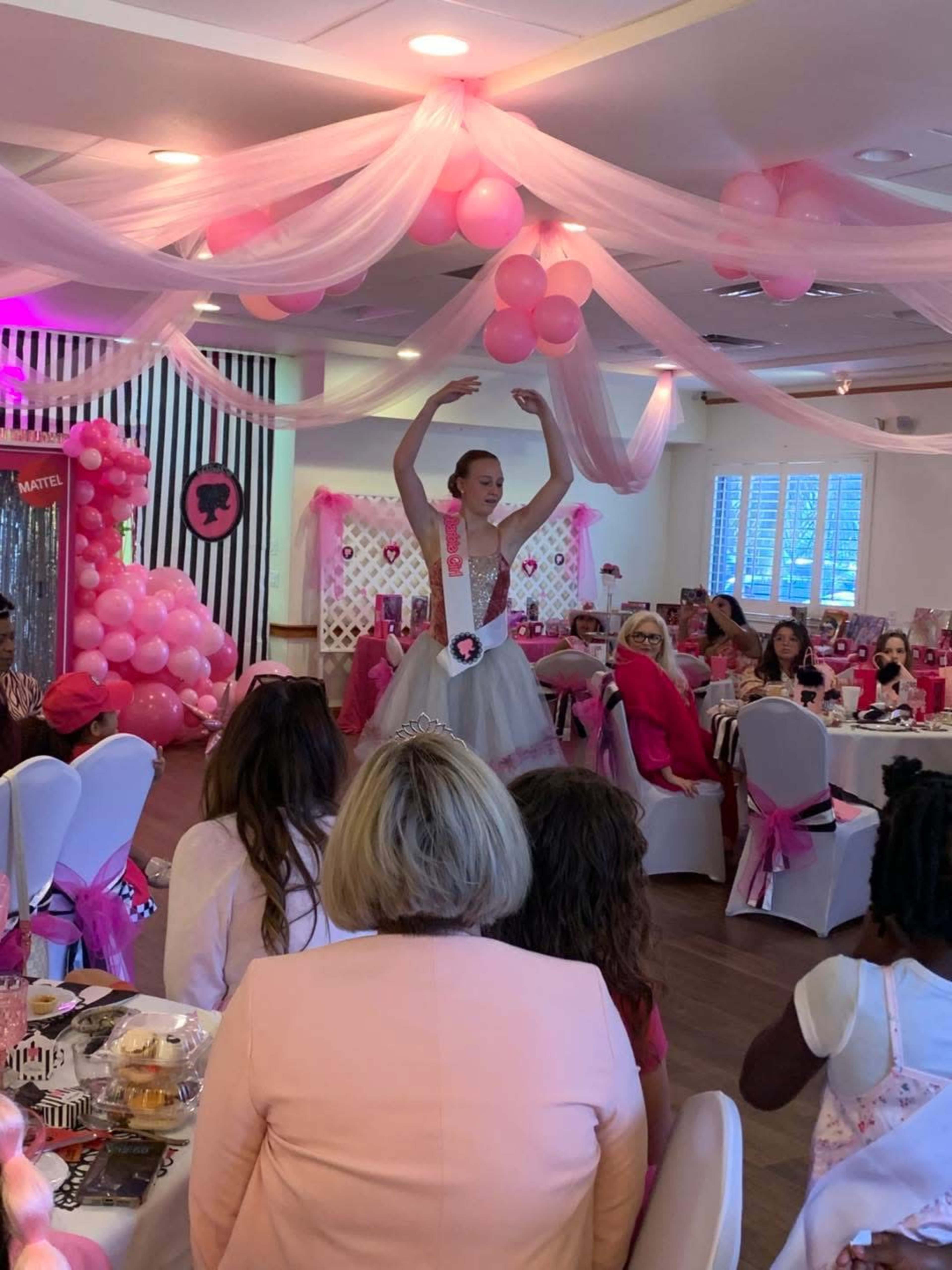 A performer in a tutu dances in front of an audience at a pink-themed event decorated with balloons and table settings.