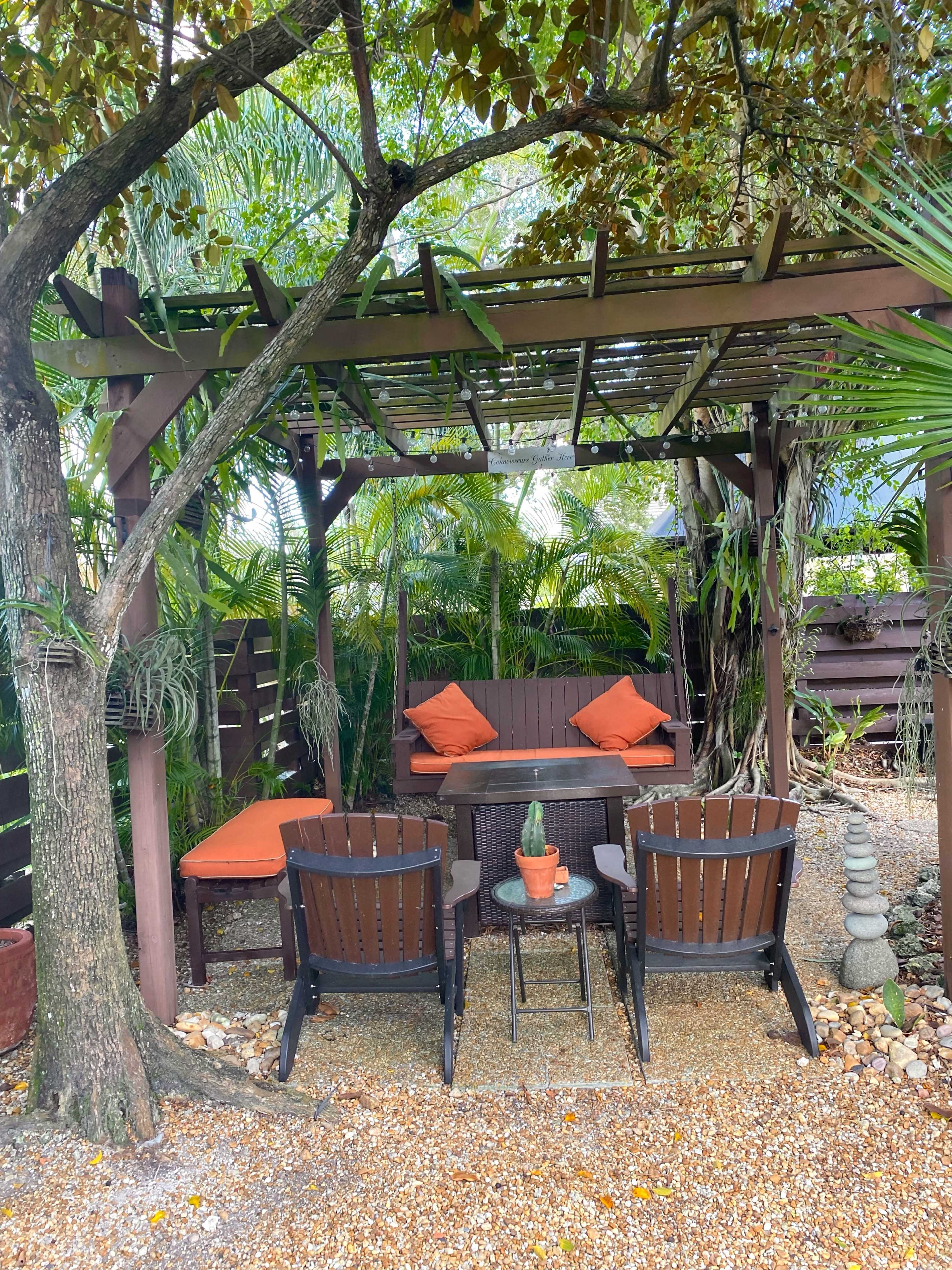 A shaded outdoor seating area features two wooden chairs, a small table with a potted plant, and cushions on a sofa surrounded by tropical greenery.