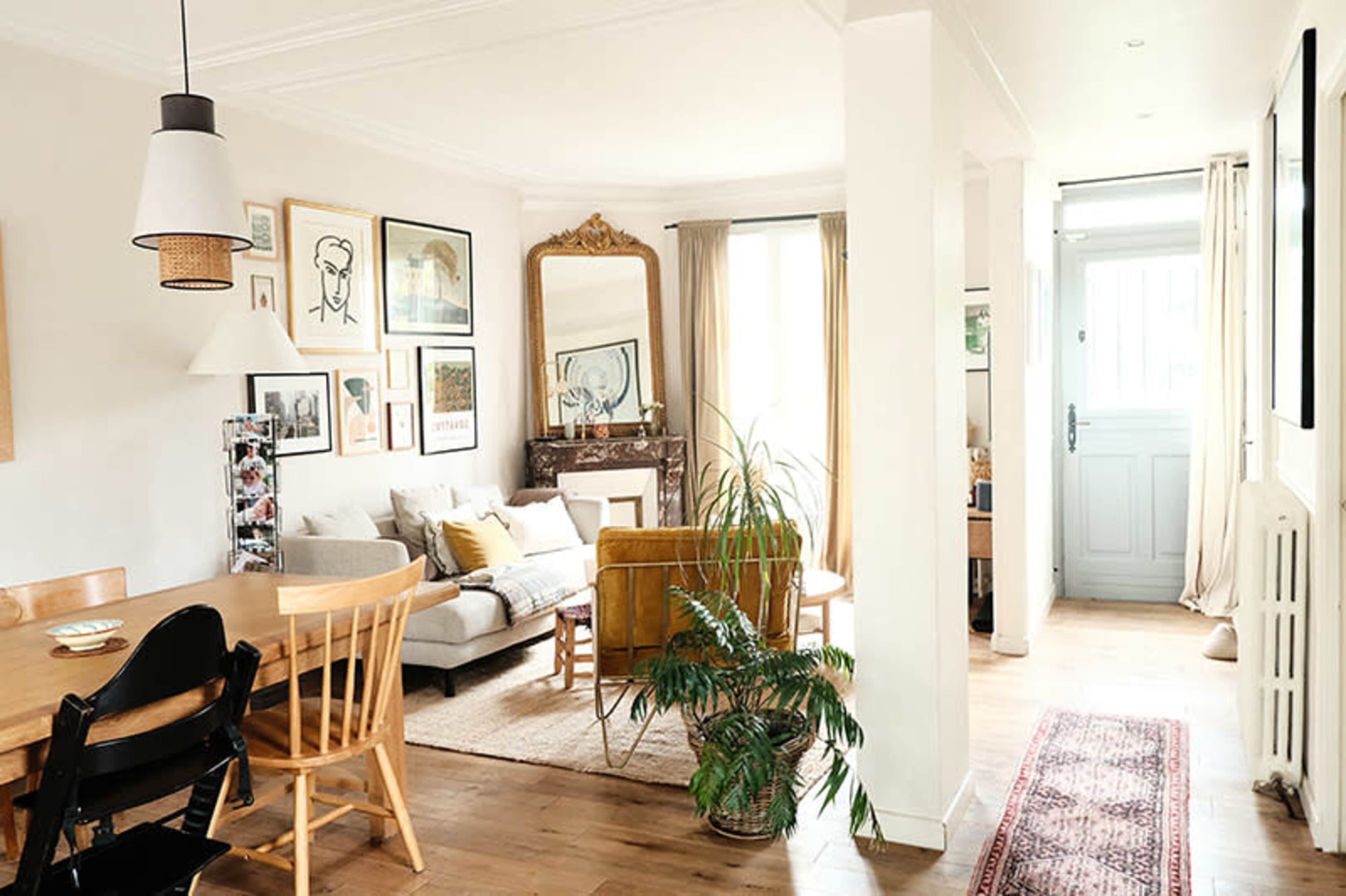 The image shows a bright, open living space featuring a dining area, seating area with a couch and chair, and a doorway leading to an entrance.