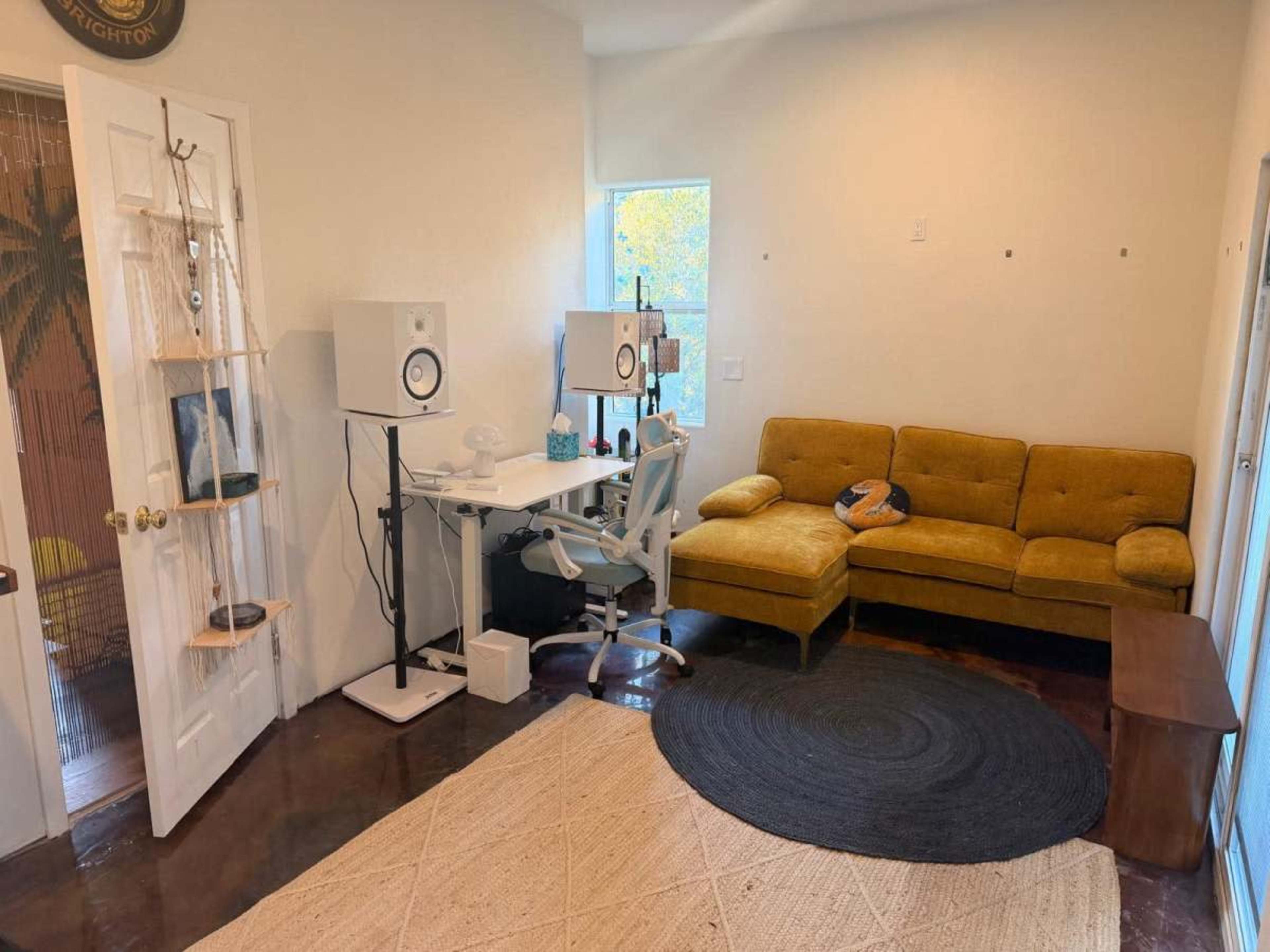 The image shows a small room with a yellow sectional couch, a desk with speakers, and a round black rug on a polished floor.