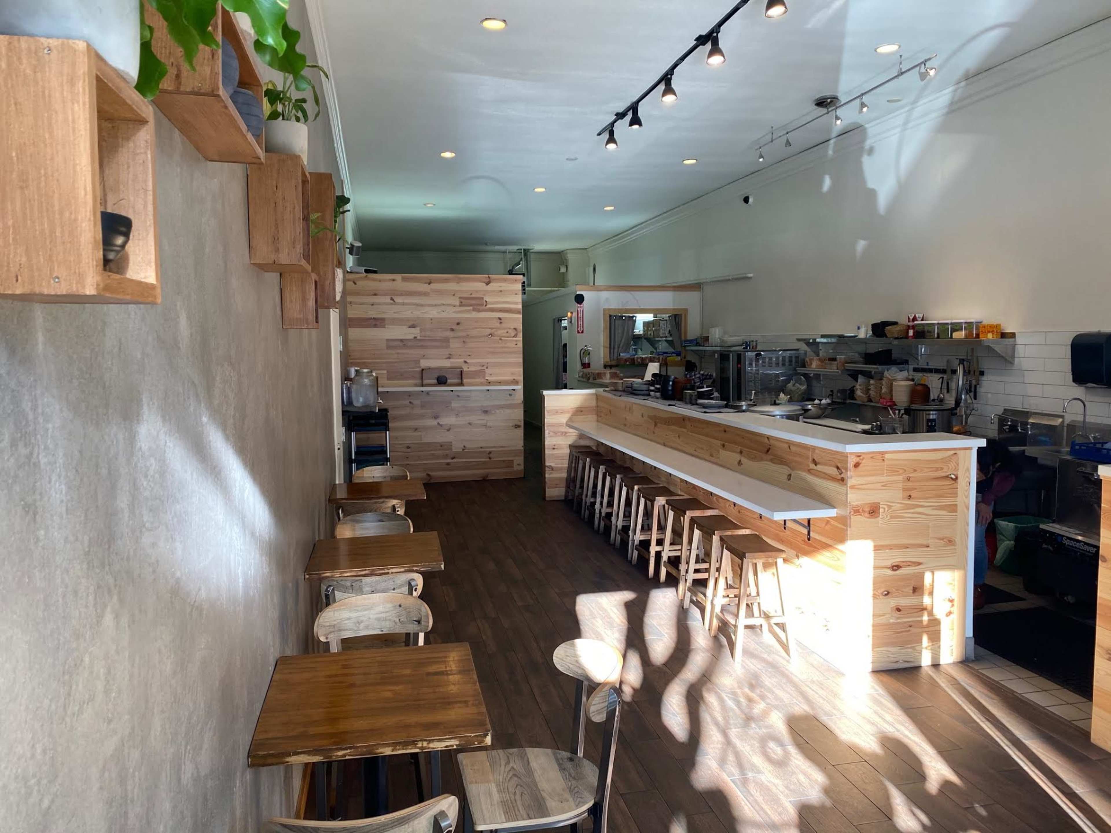 The interior of a minimalist café features wooden furniture and paneling, with sunlight streaming in through the windows.
