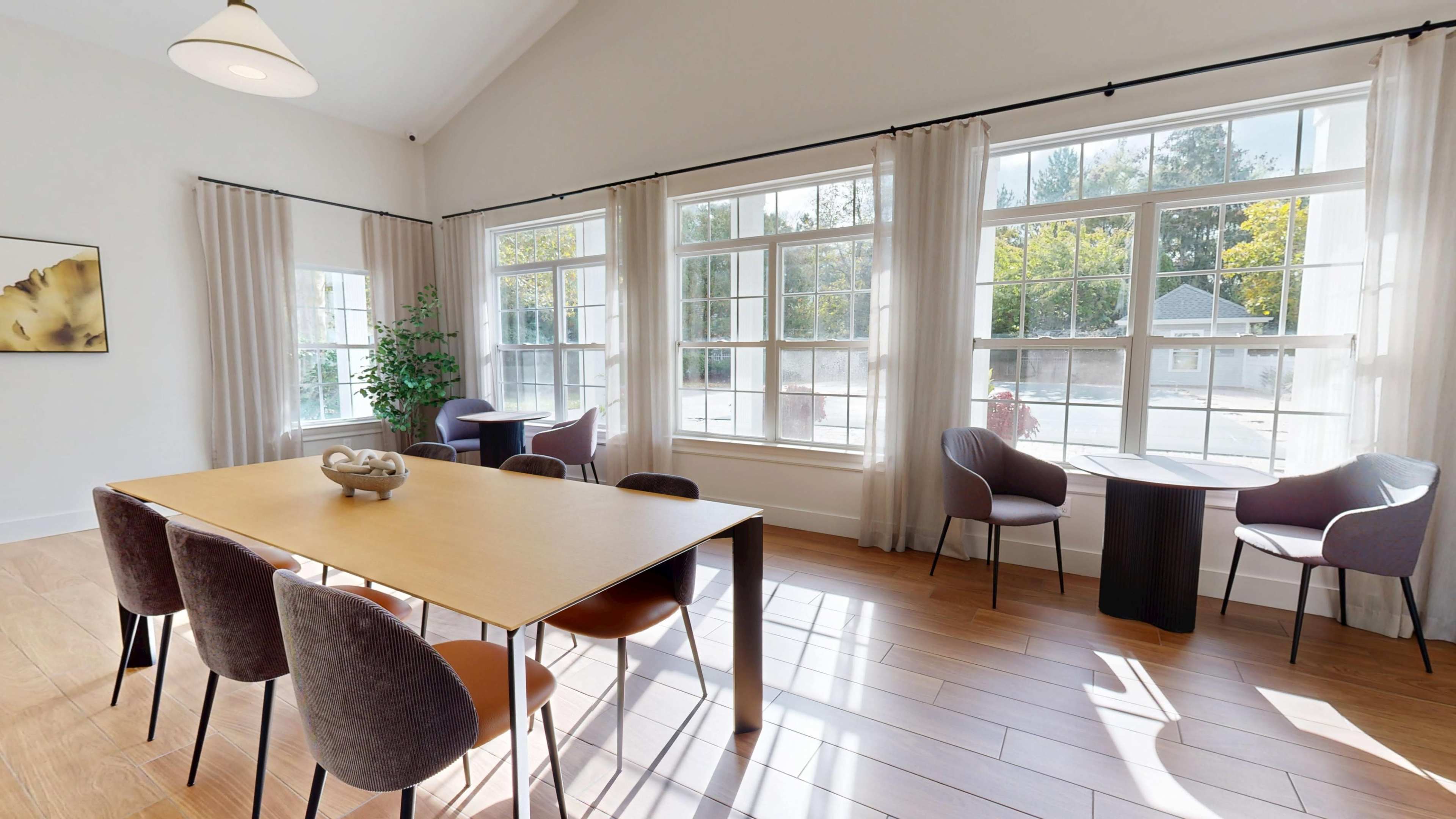 The image shows a well-lit dining area with a wooden table surrounded by chairs, large windows with sheer curtains, and a view of greenery outside.