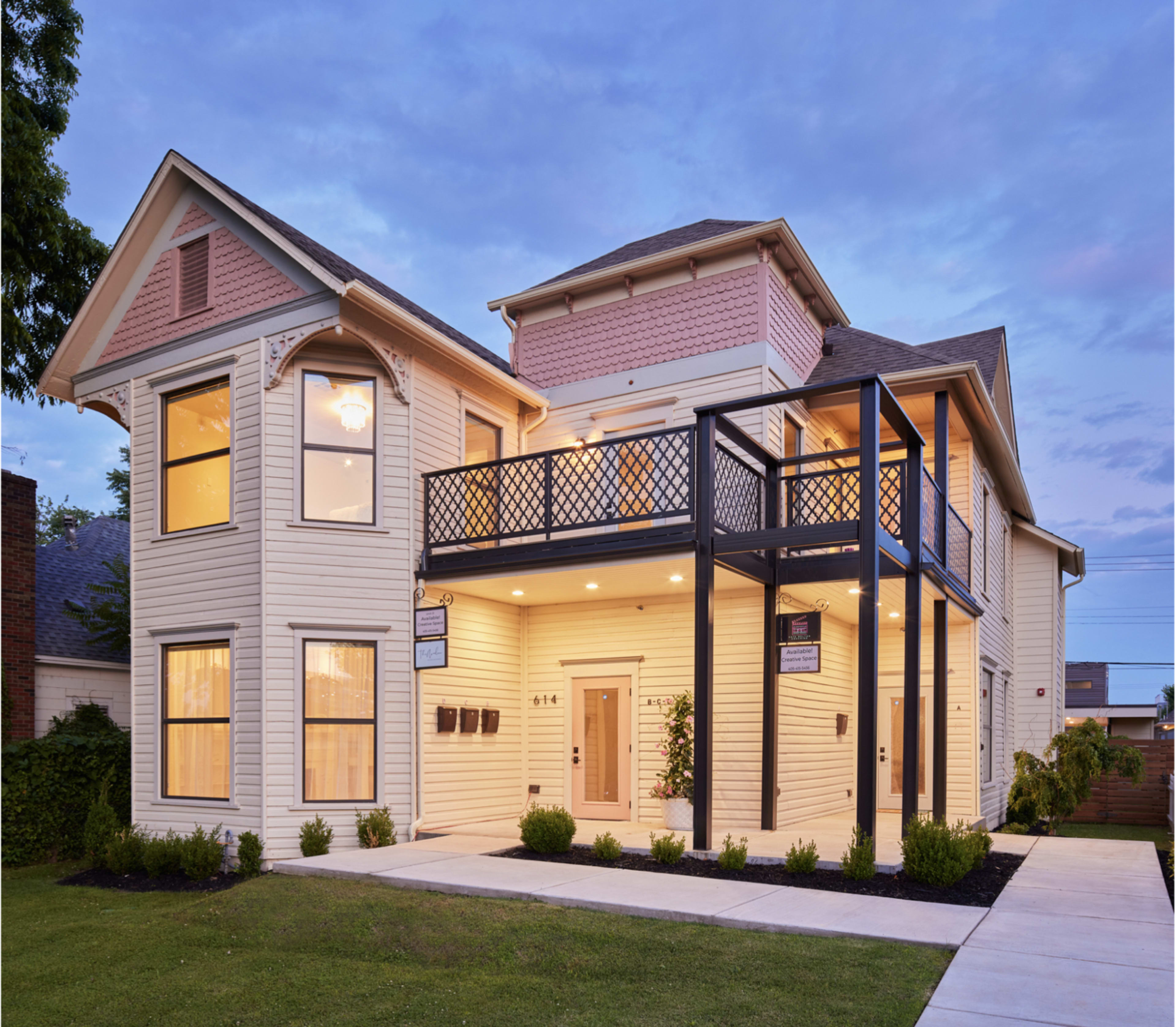 A two-story, light-colored house with a front porch, large windows, and a black balcony railing.
