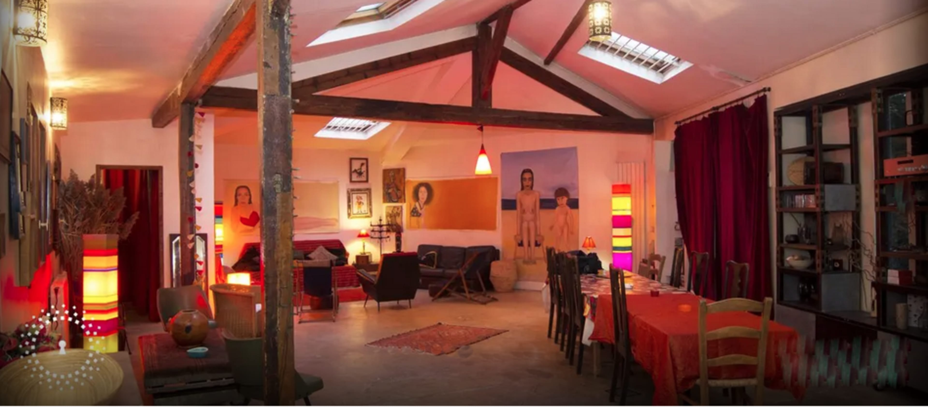 A spacious room features a high ceiling supported by wooden beams, with dining and seating areas decorated in a mix of vibrant colors and artistic wall murals.