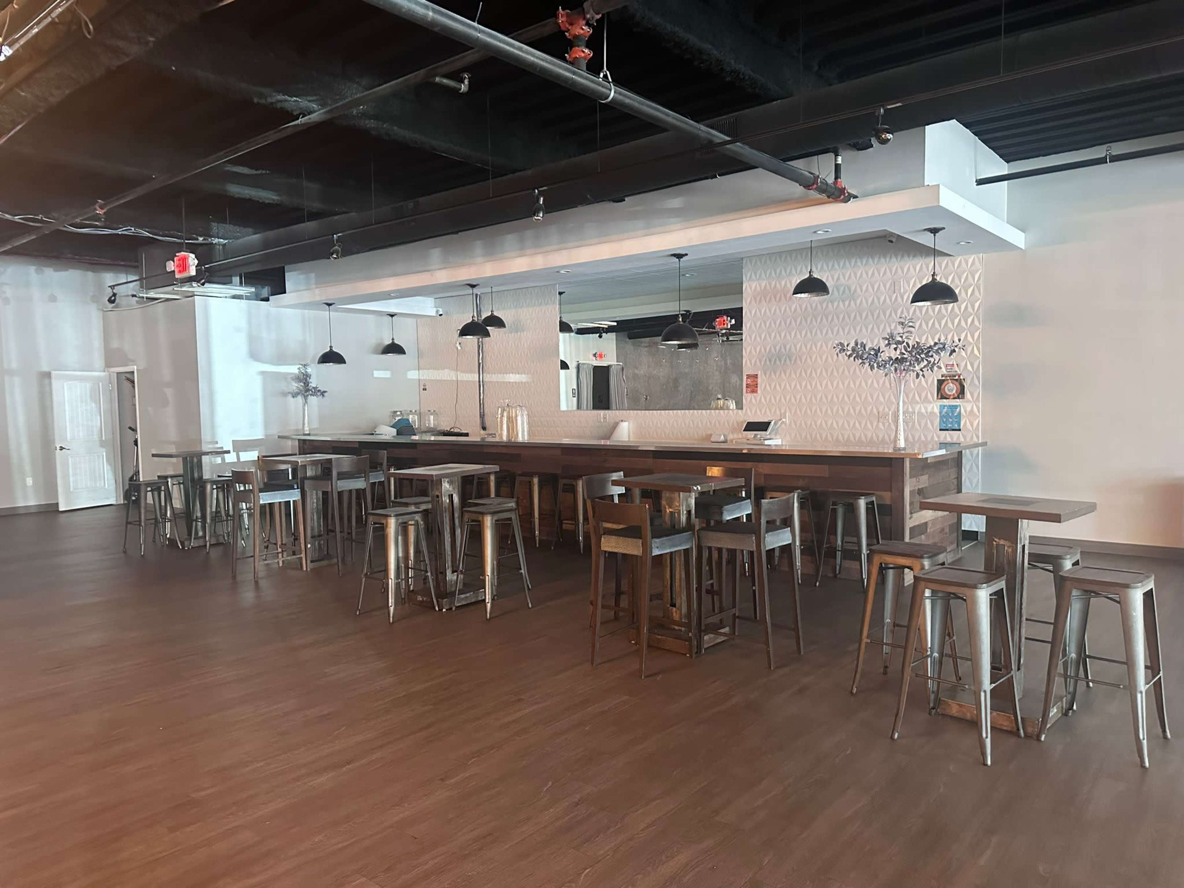 The image shows a spacious, empty bar area with wooden and metal seating, illuminated by overhead pendant lights.