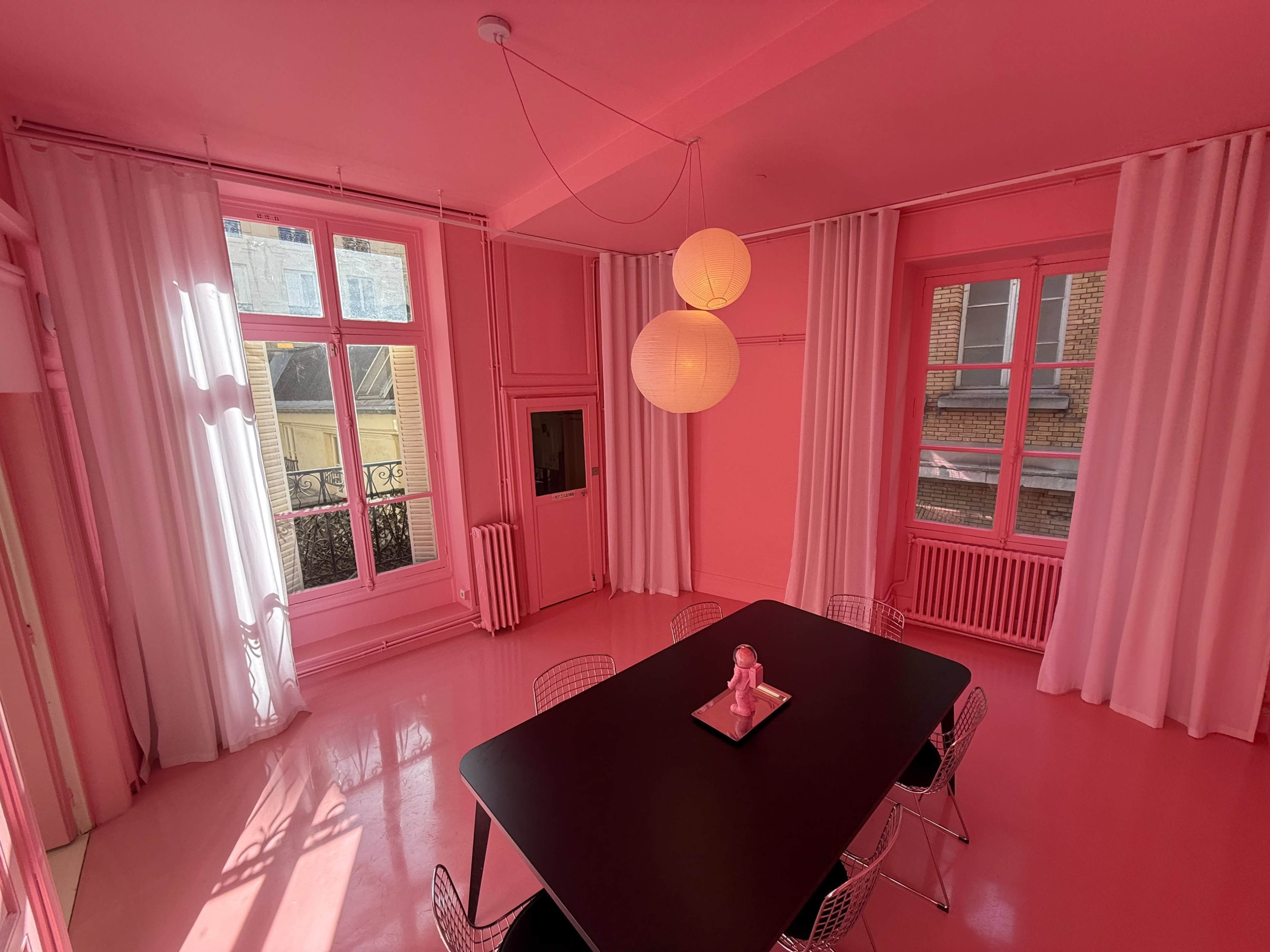 The image features a brightly lit room with pink walls, a pink floor, and a black dining table surrounded by wire chairs, illuminated by two pendant lamps.