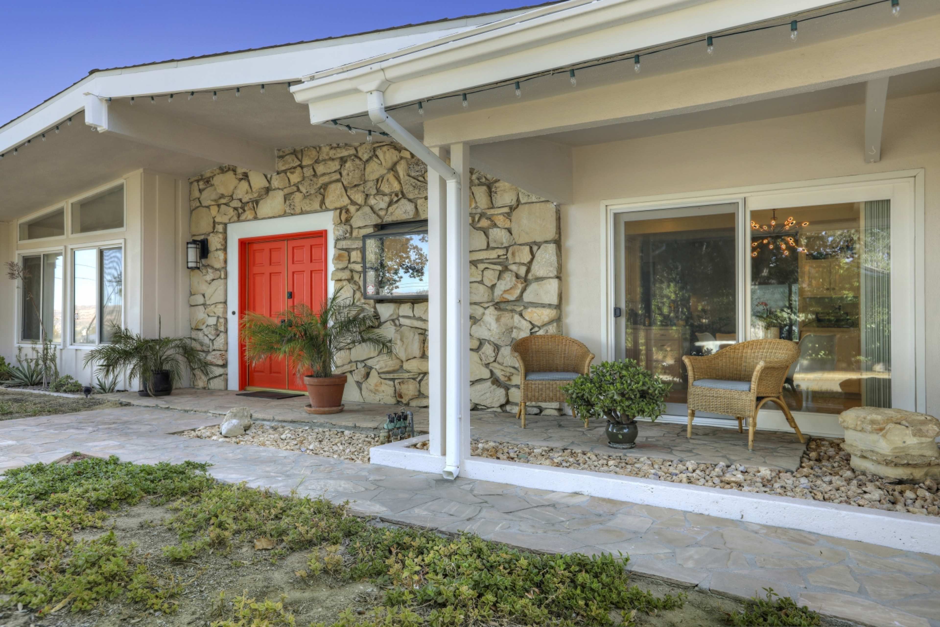 A front porch with a stone wall, a bright red door, two wicker chairs, and decorative plants.