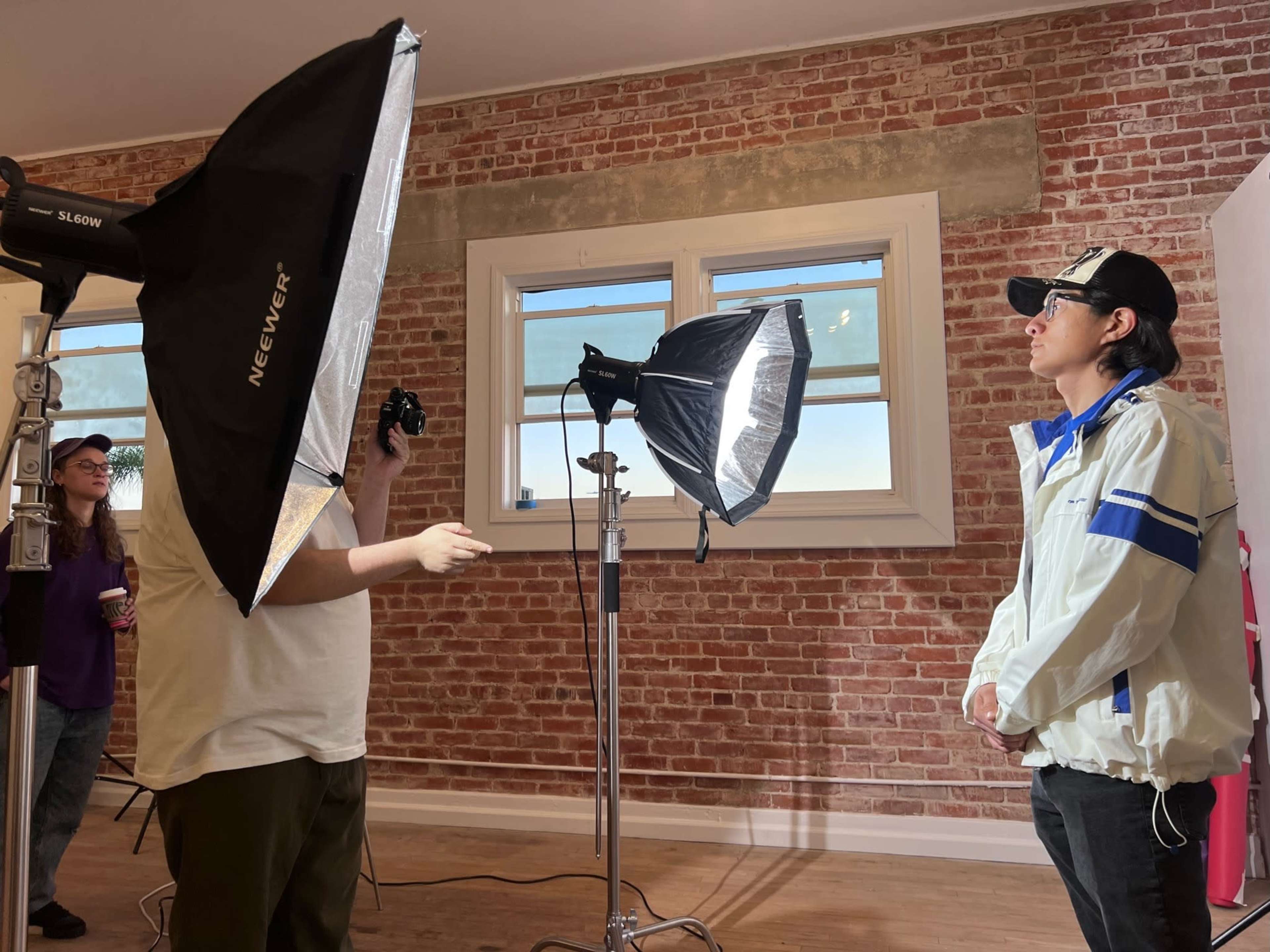 A person in a white jacket stands in front of two studio lights while another person operates a camera in a room with exposed brick walls.