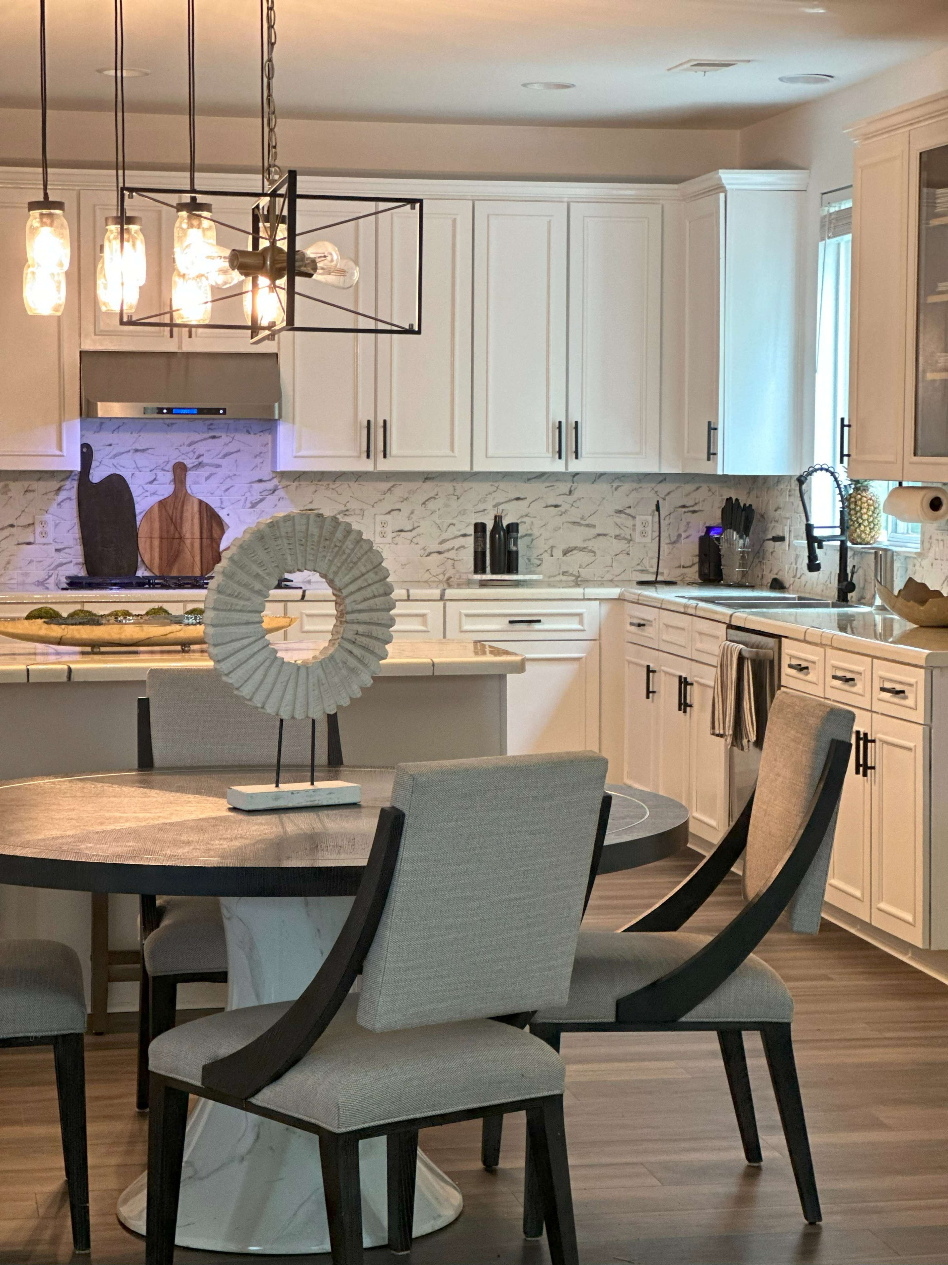 A modern kitchen features white cabinetry, a marble-topped table, and a decorative centerpiece, illuminated by pendant lighting.