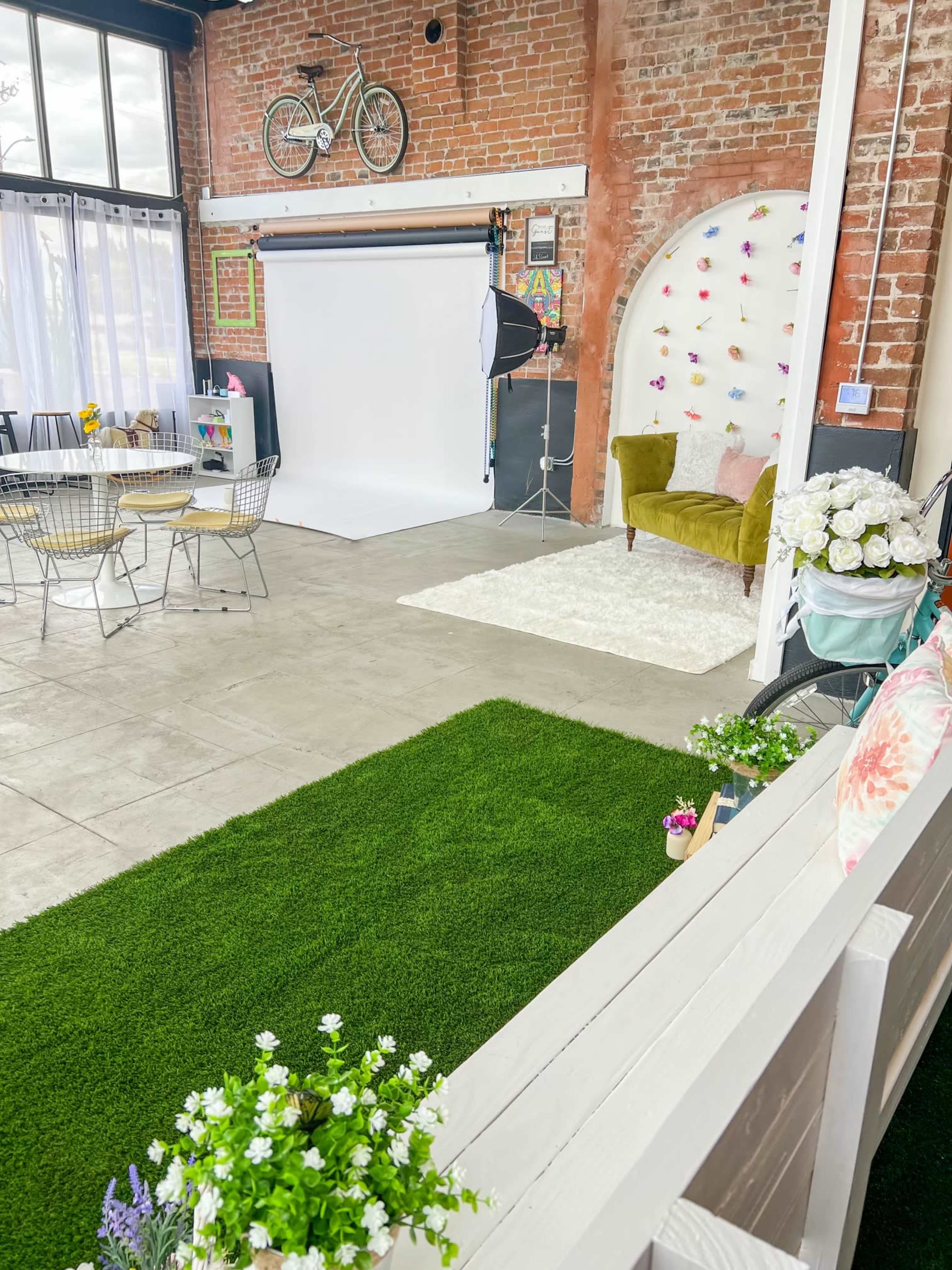 The scene features a photography studio with a white backdrop, a green carpet, seating areas, and decorative elements like flowers and a bicycle on exposed brick walls.