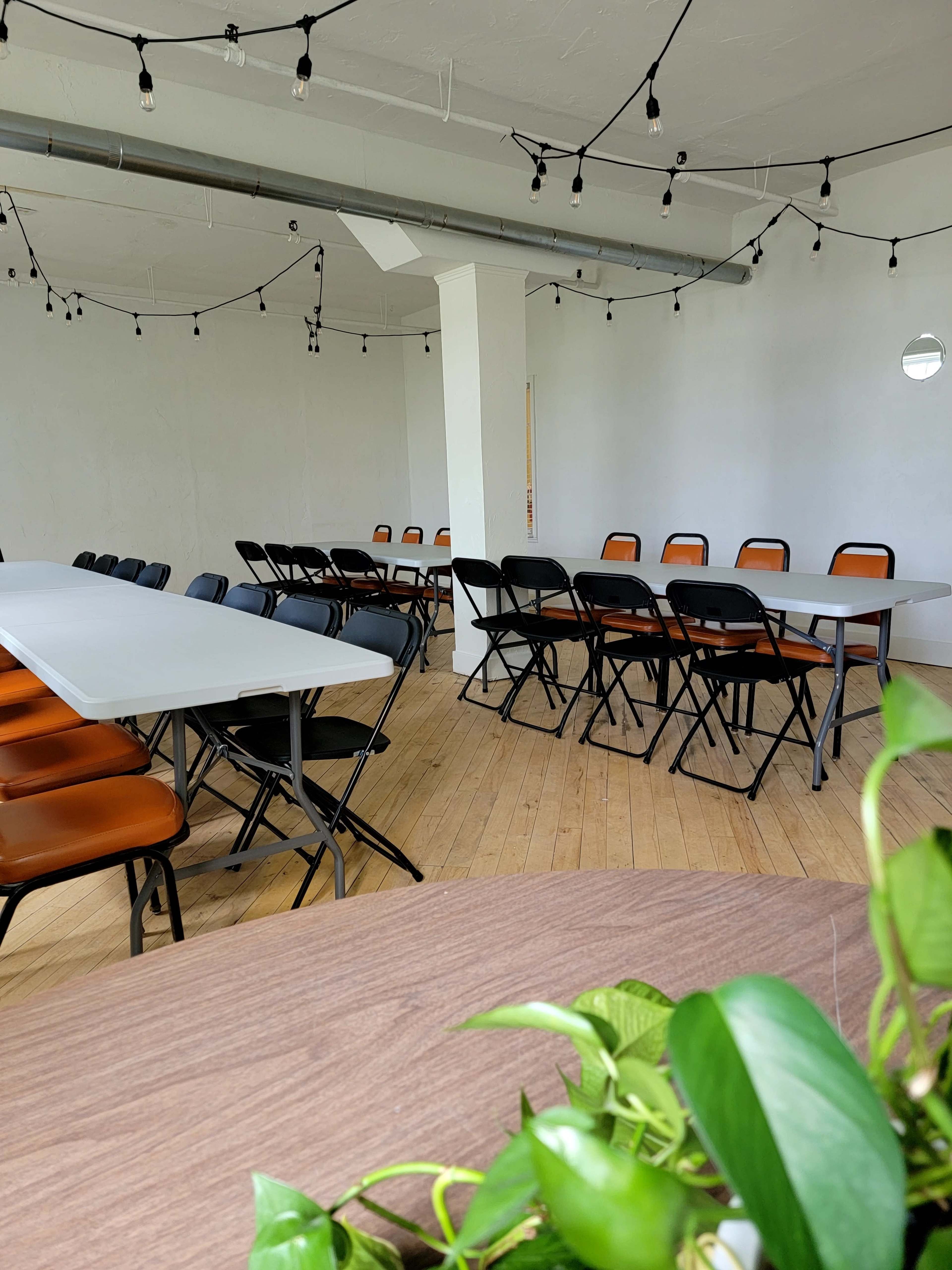 A spacious room features several tables arranged in a U-shape, surrounded by black and orange folding chairs, with exposed light bulbs hanging from the ceiling.