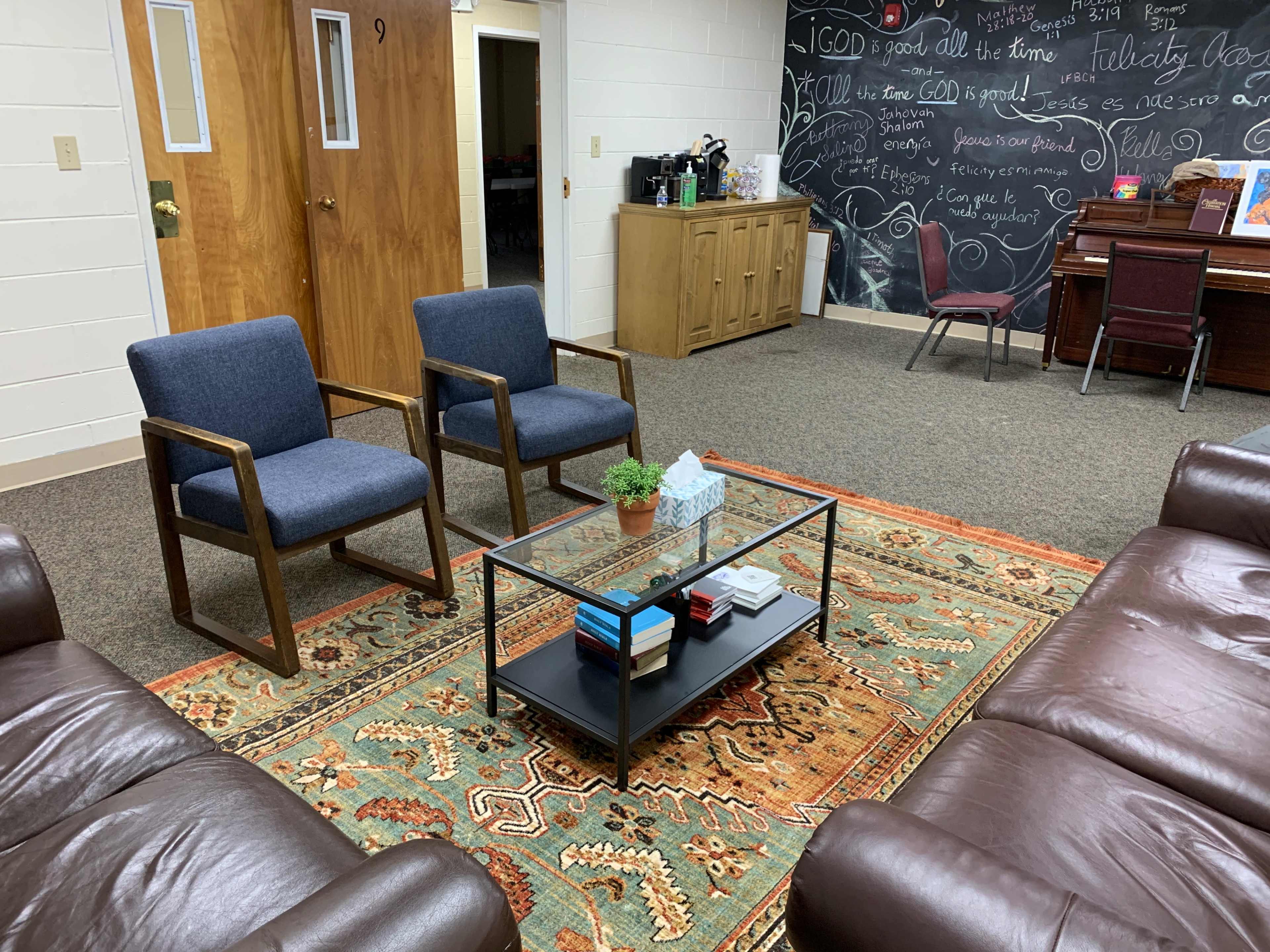 A cozy lounge area with two blue chairs facing a glass coffee table on an ornate rug, surrounded by brown leather couches in a room with a chalkboard wall and a small kitchenette in the background.
